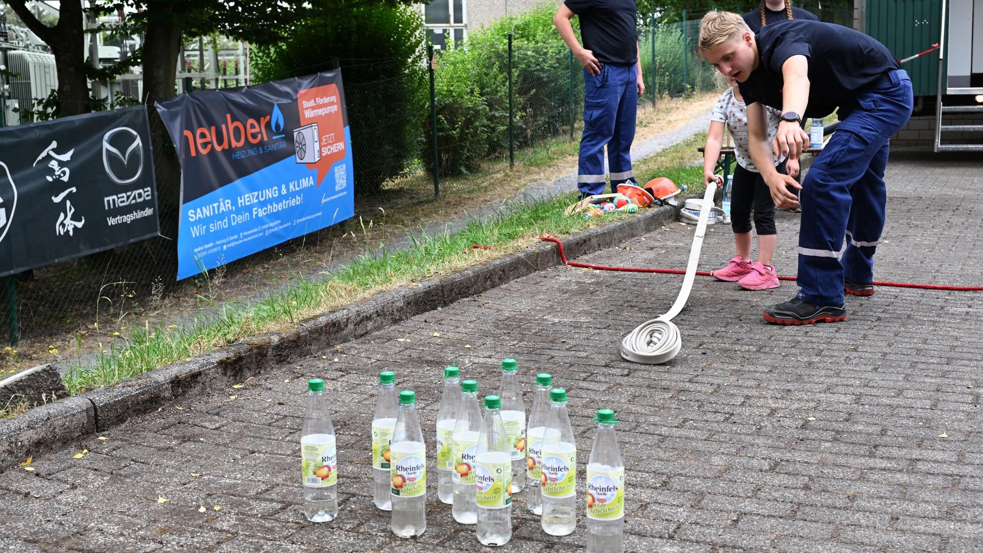 Zu sehen ist ein junger Feuerwehrmann, der einem Kind zeigt, wie man einen Feuerwehrschlauch ausrollt.
