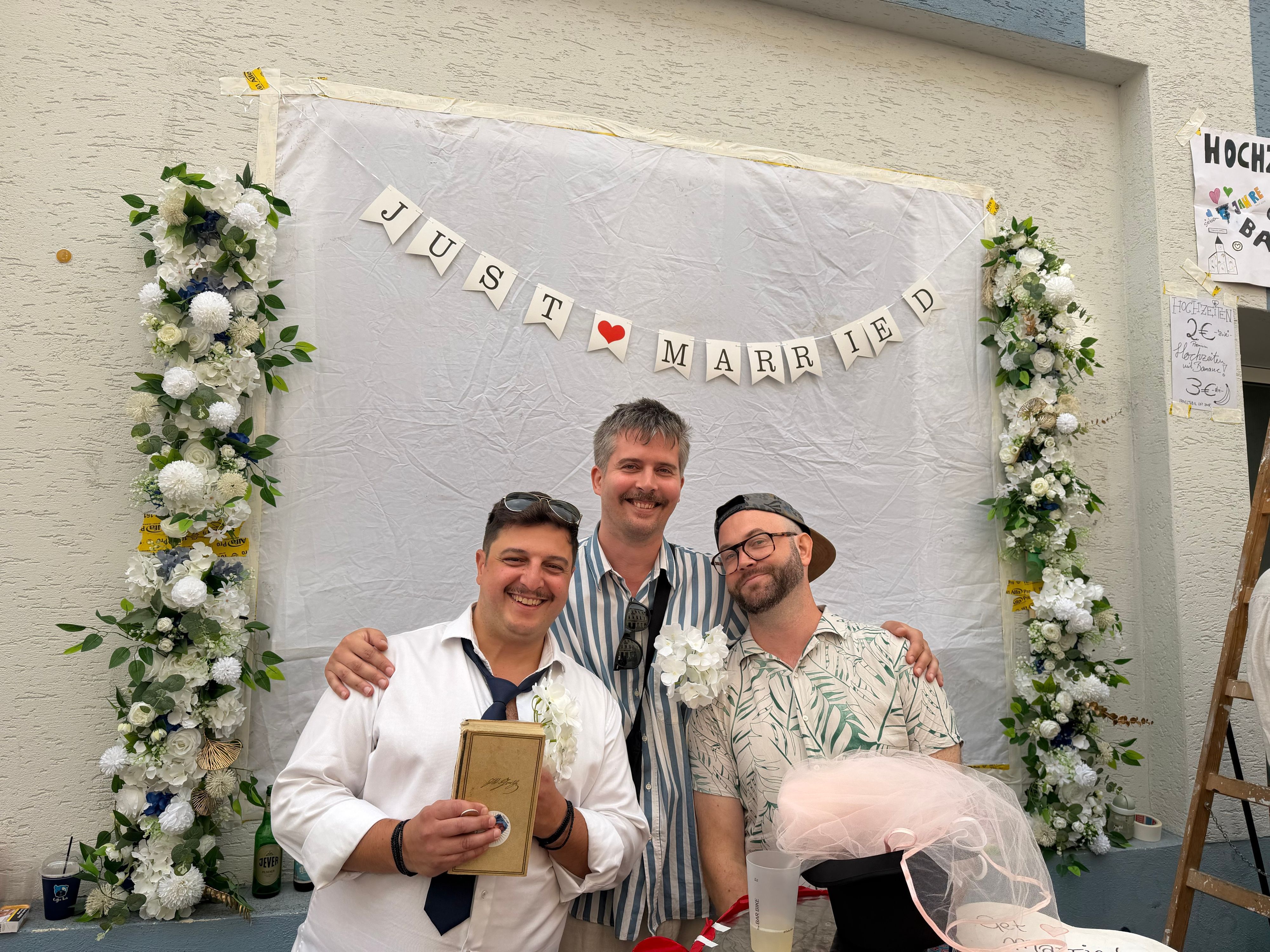 Drei Männer stehen unter einem mit Blumen geschmückten Schriftzug "Just Married".