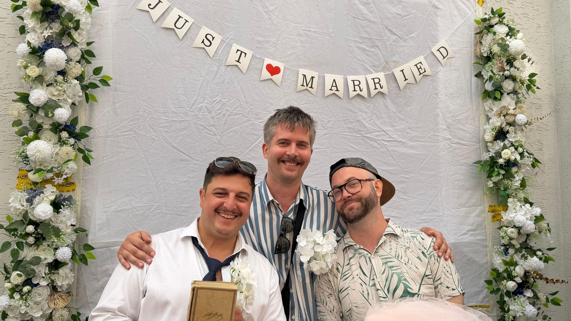 Drei Männer stehen unter einem mit Blumen geschmückten Schriftzug "Just Married".