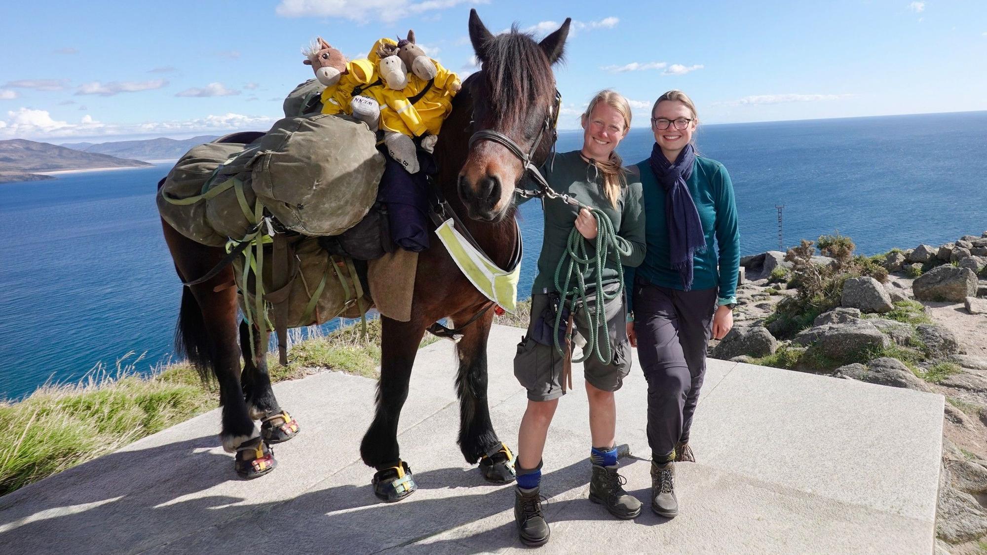Zu sehen ist ein Pferd mit zwei Frauen. Sie stehen in der Nähe einer Klippe mit dem Rücken zum Meer.