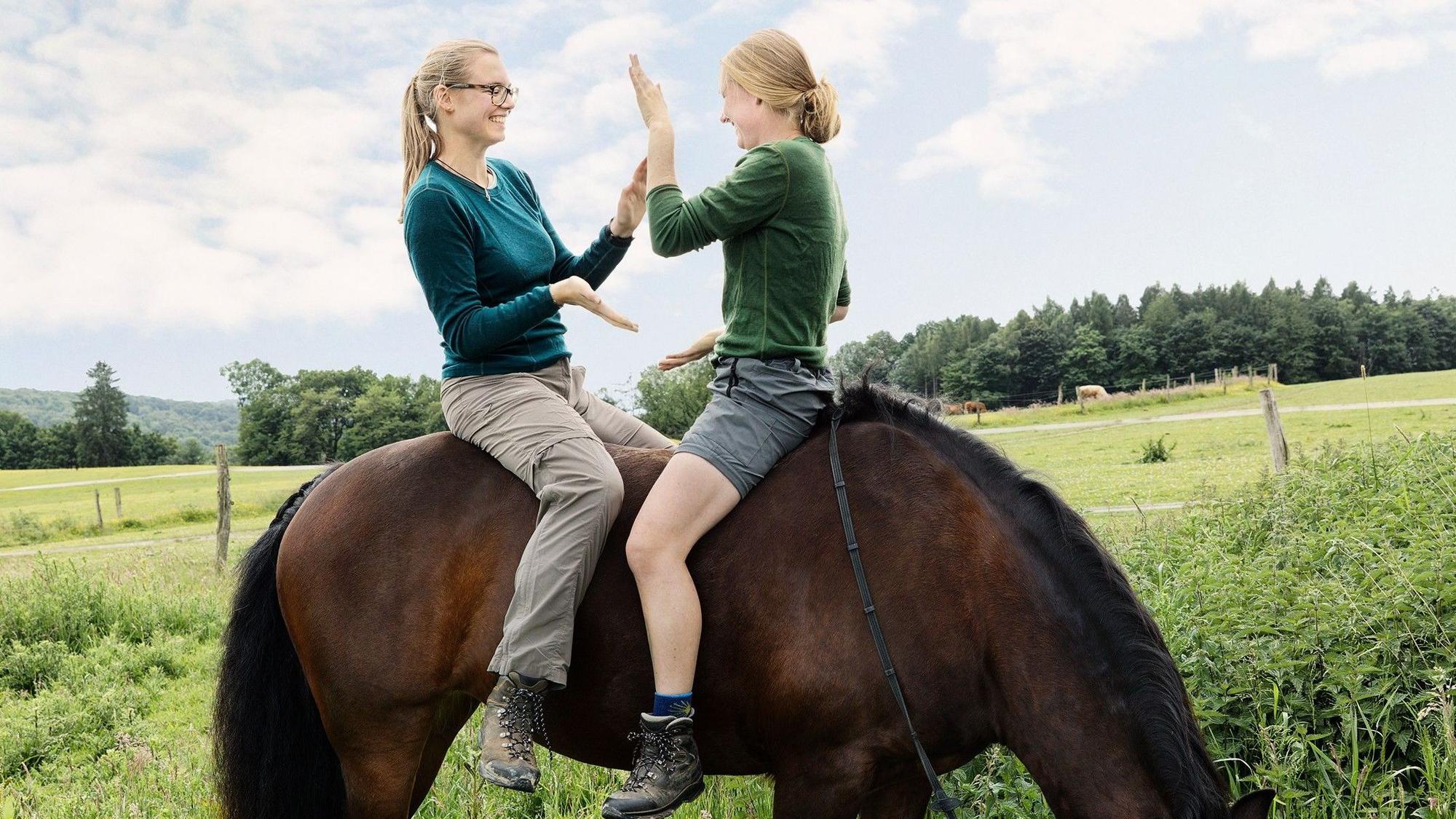 Zwei junge Frauen sitzen auf dem Rücken eines Pferdes und klatschen sich gegenseitig ab.