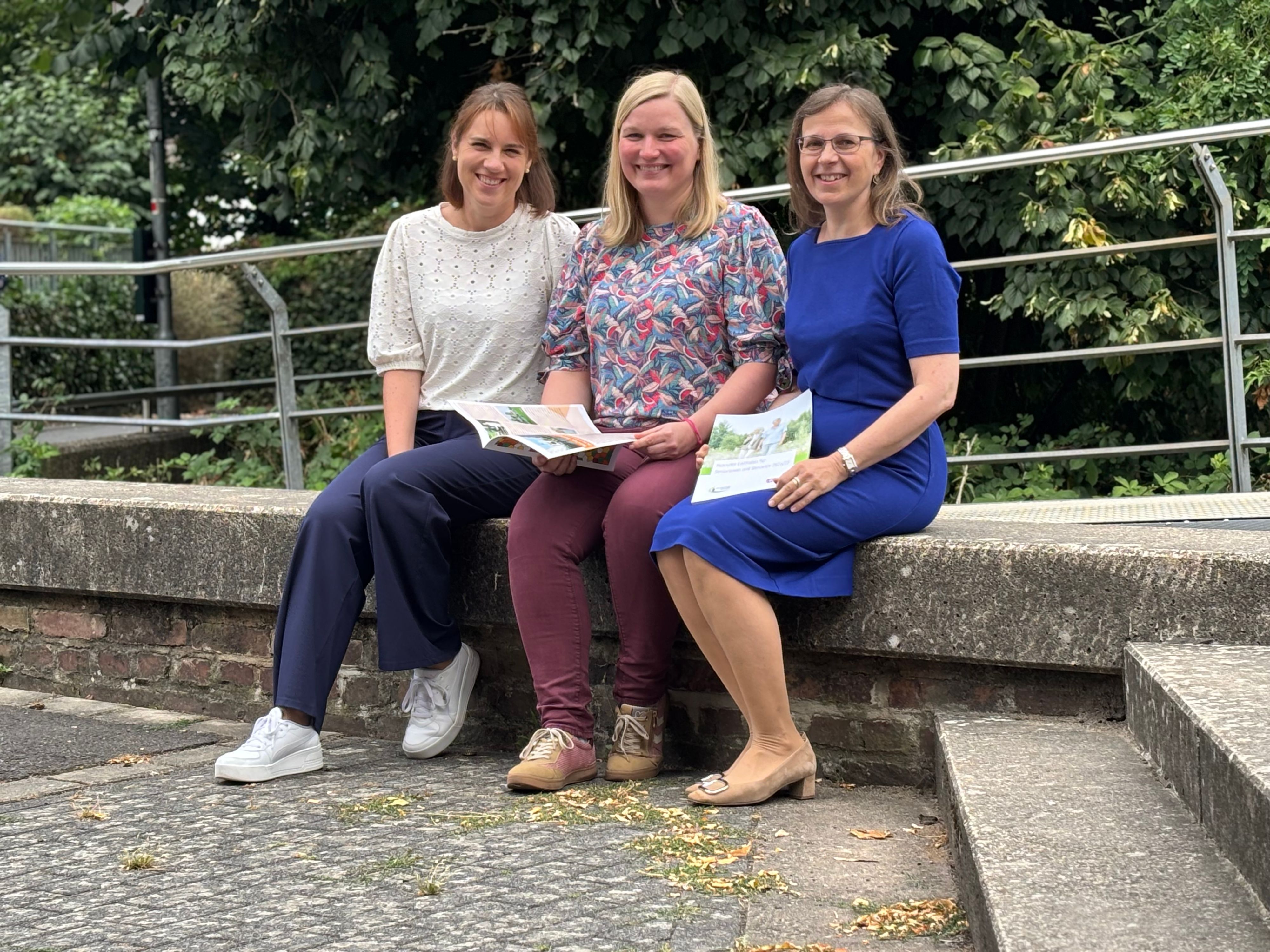 Die Obengenannten sitzen draußen auf einer Mauer. Lena Bleich und Doris Hofmann halten Informationsmaterial für ältere Menschen in den Händen.