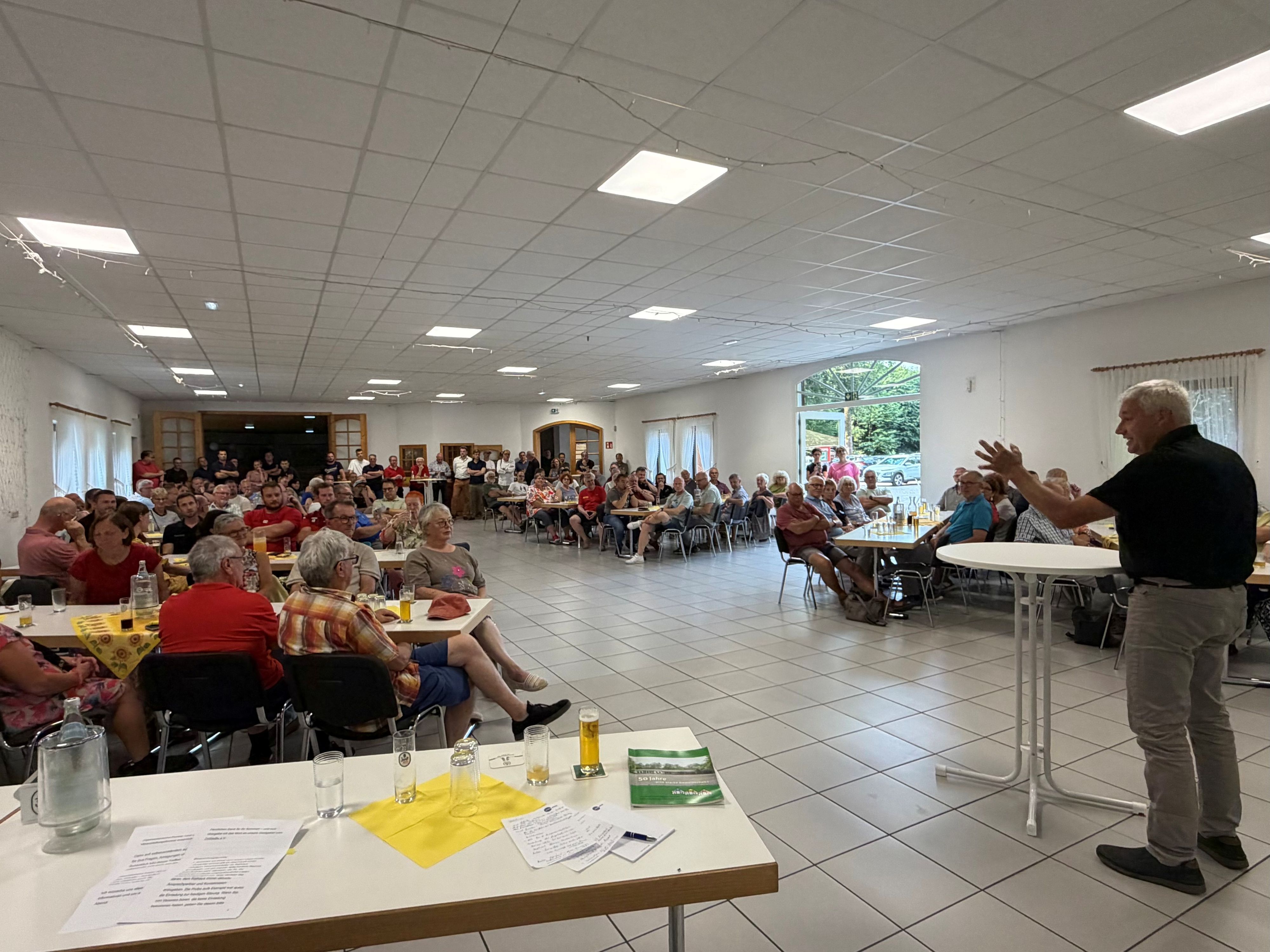 Ein Mann am Stehtisch begrüßt sitzende Gäste.