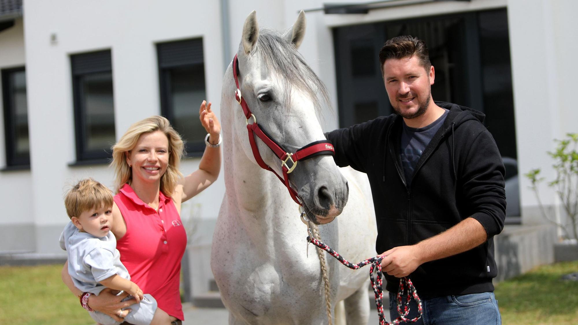 Ein Mann und eine Frau neben einem weißen Pferd; die Frau hat ein kleines Kind auf dem Arm.
