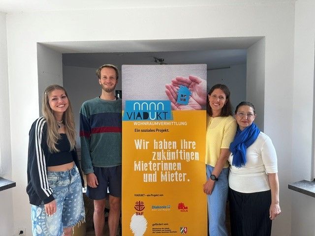 Drei Frauen und ein Mann stehen links und rechts von einem Aufsteller, auf dem steht „Wir haben Ihre zukünftigen Mieterinnen und Mieter“.