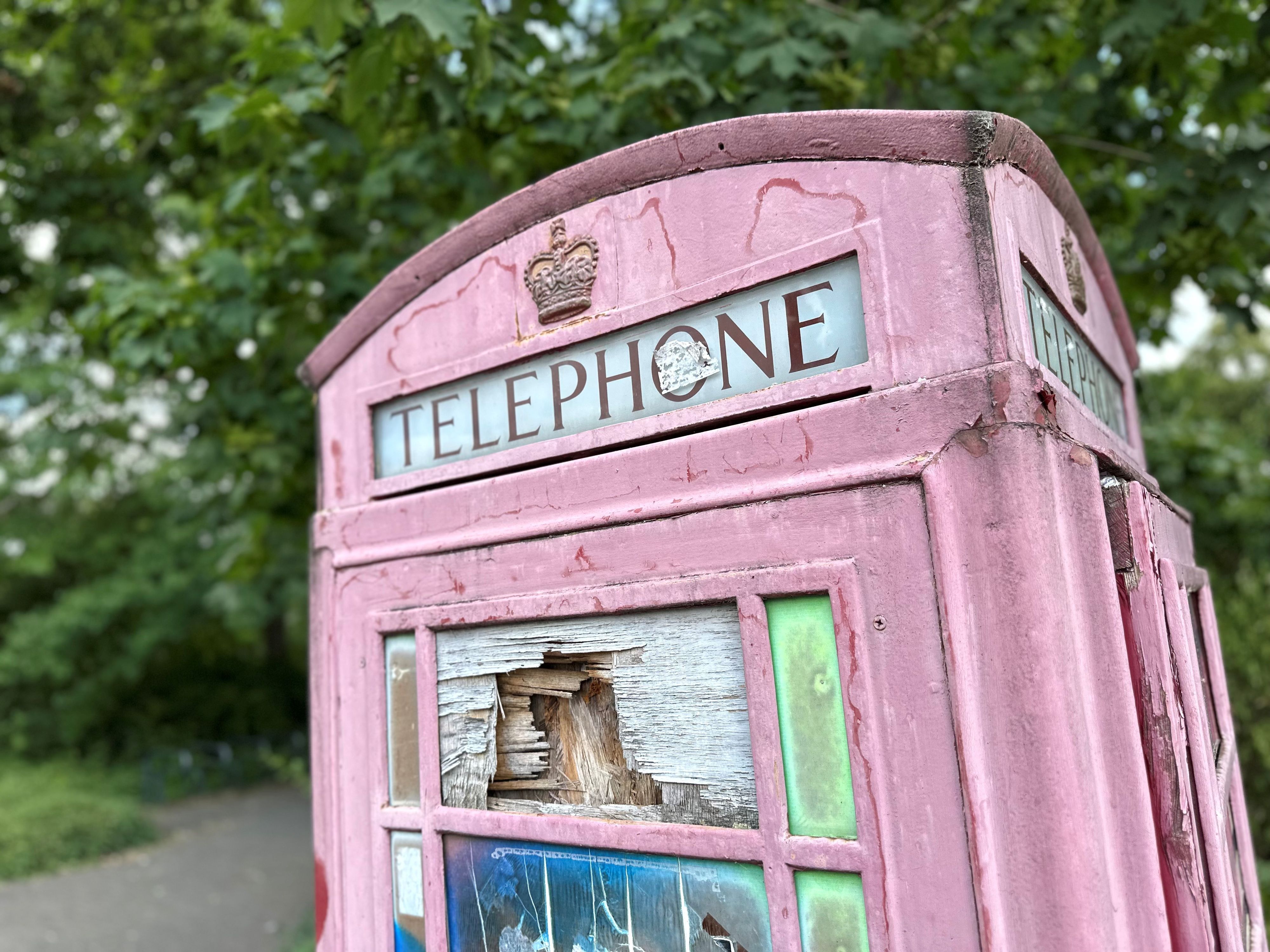 Eine rote Telefonzelle steht auf einem Platz, dahinter sind Beete und Bäume zu sehen.