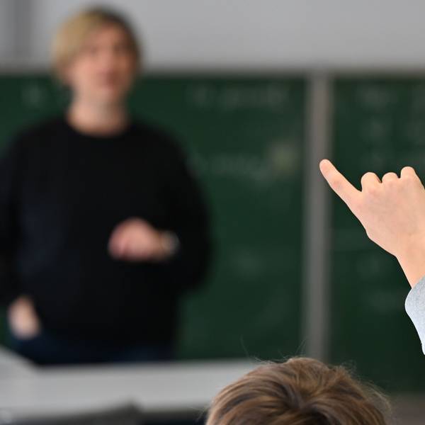 Ein Schüler zeigt im Klassenraum auf, um sich zu melden und blickt zur Lehrerin im Hintergrund. (Symbolfoto)