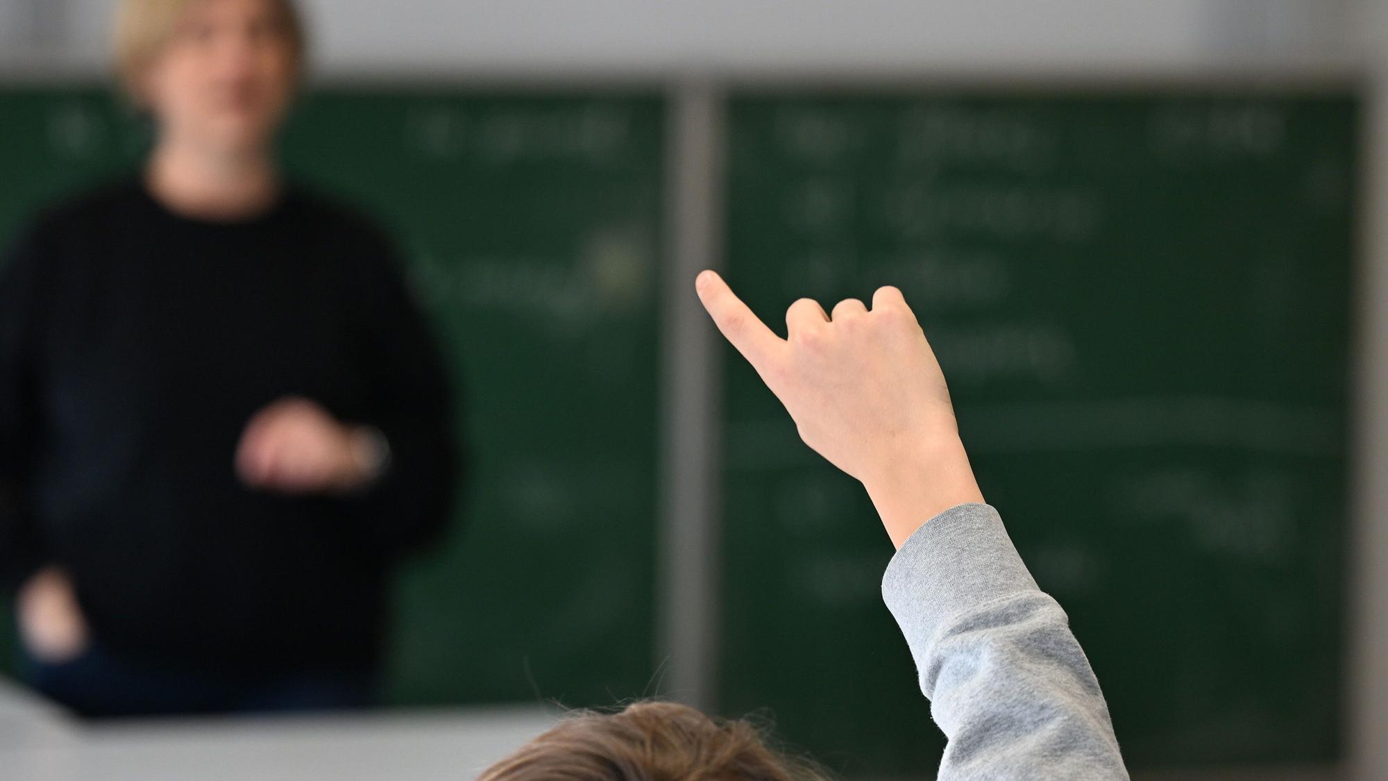 Ein Schüler zeigt im Klassenraum auf, um sich zu melden und blickt zur Lehrerin im Hintergrund. (Symbolfoto)