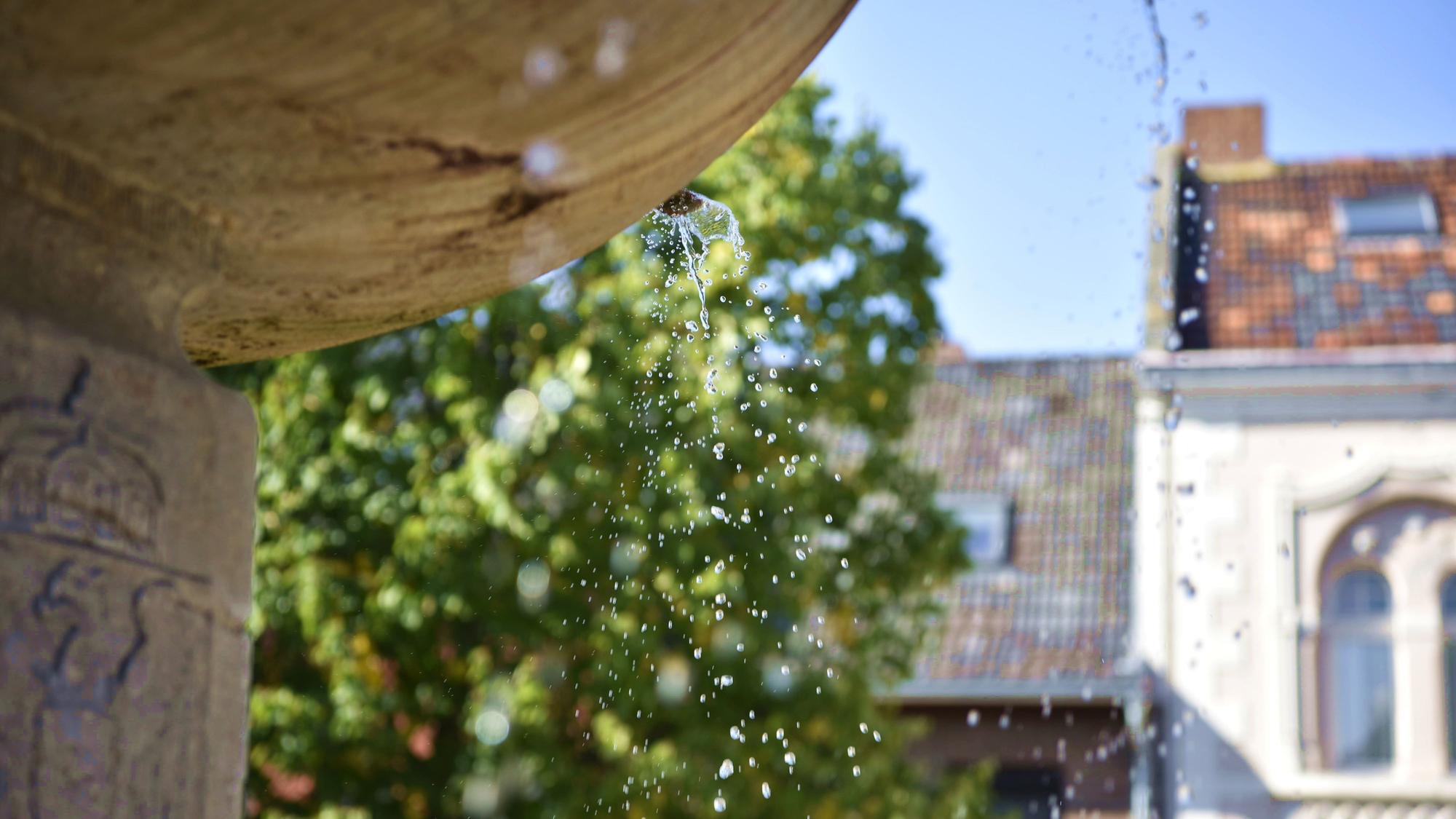 Aus einem Brunnen tropft Wasser, im Hintergrund sind Häuser und Bäume zu erkennen.