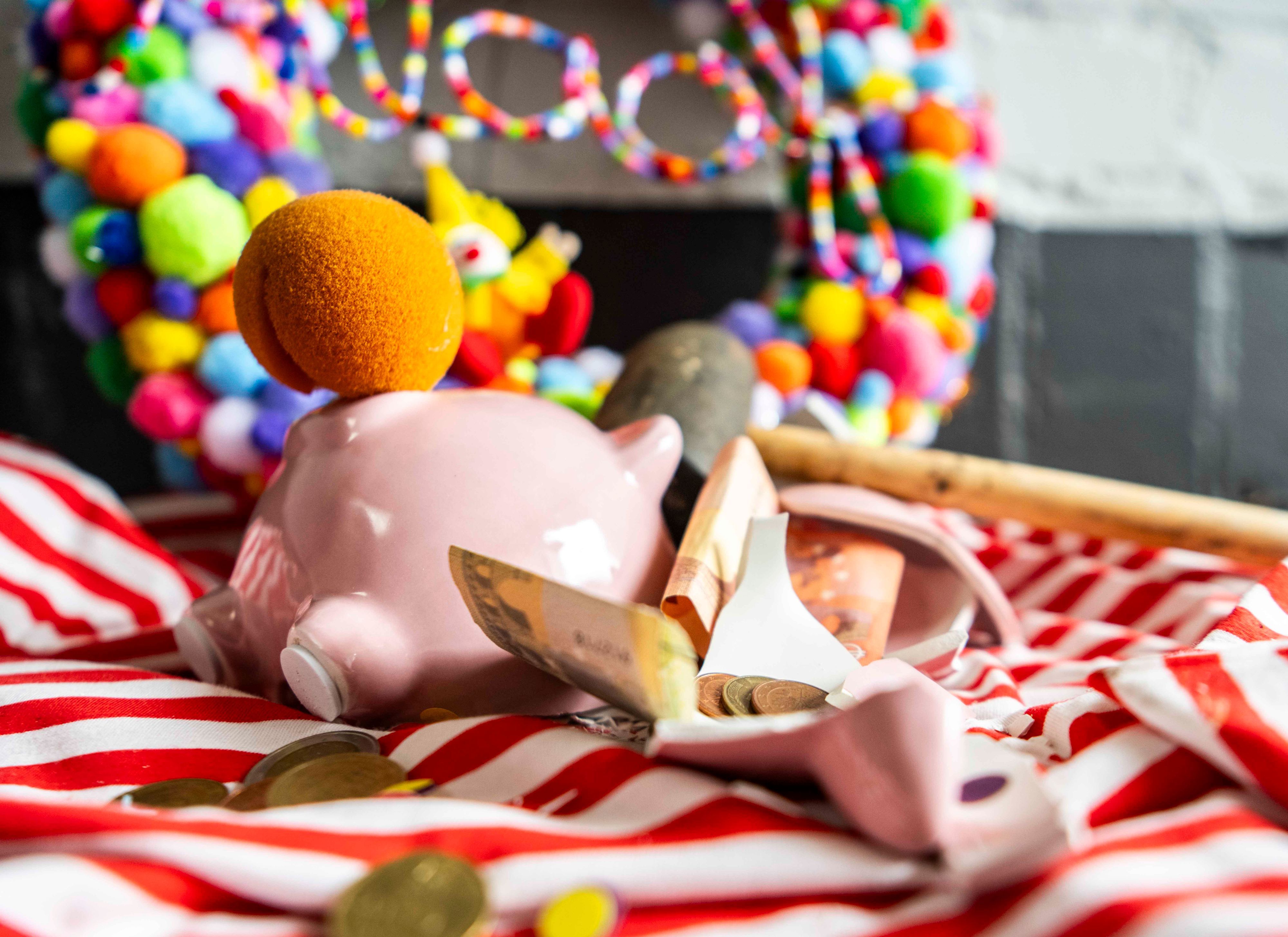 Das Bild zeigt ein zerschlagenes Sparschwein, das eine Clownsnase anhat. Auch Geld und ein Karnevalskranz sind zu sehen.