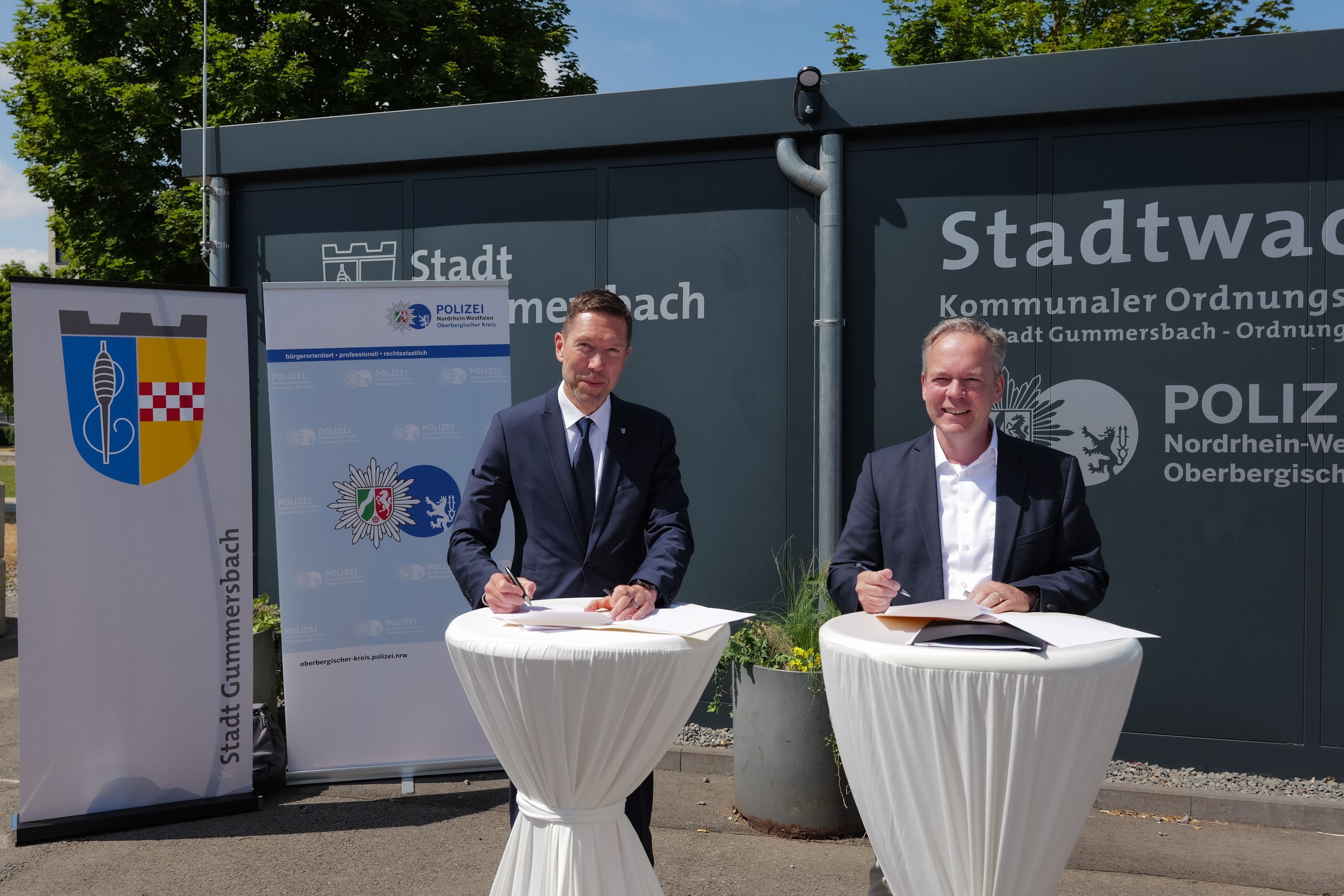 Zwei Männer stehen an Tischen und unterschreiben eine Kooperationsvereinbarung.