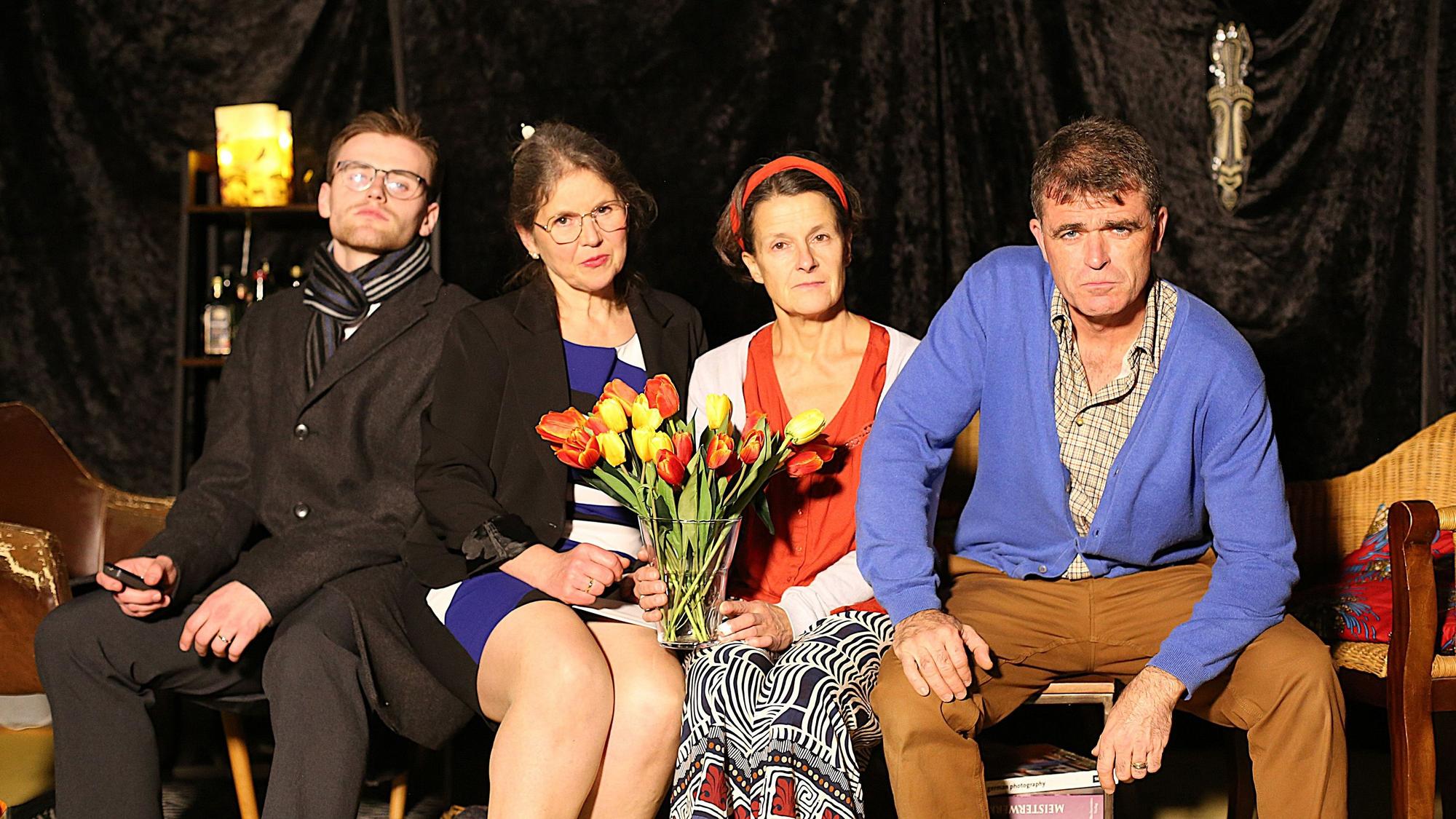 Zwei Schauspielerinnen und zwei Schauspieler sitzen nebeneinander. Eine Frau hält einen Strauß künstlicher Blumen.