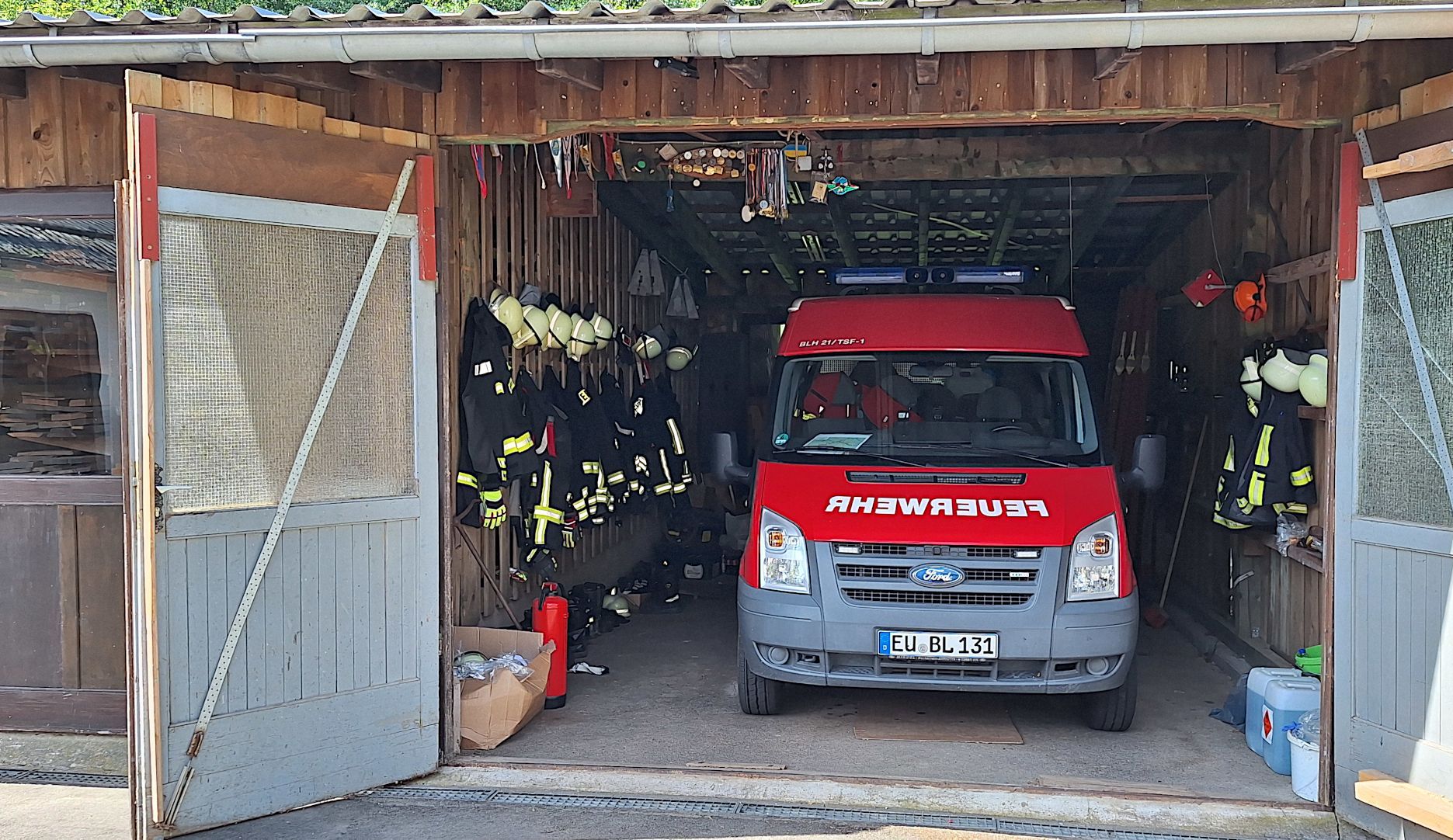 Ein rotes Feuerwehrauto steht in einm Holzschuppen. Links und rechts daneben hängt Feuerwehrkleidung.