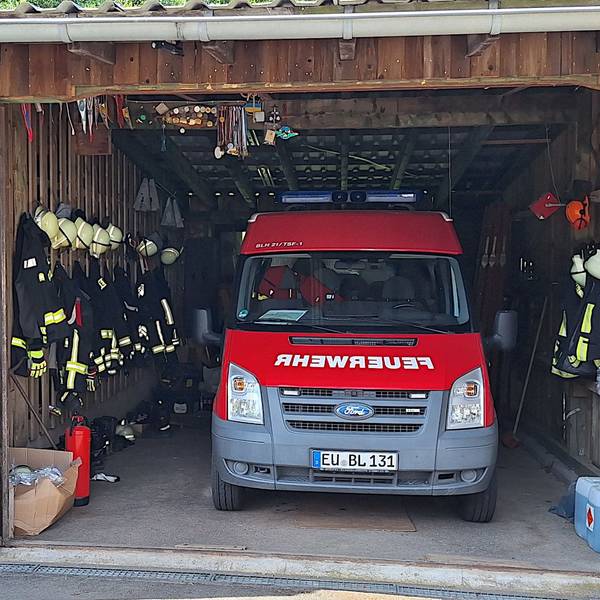 Ein rotes Feuerwehrauto steht in einm Holzschuppen. Links und rechts daneben hängt Feuerwehrkleidung.