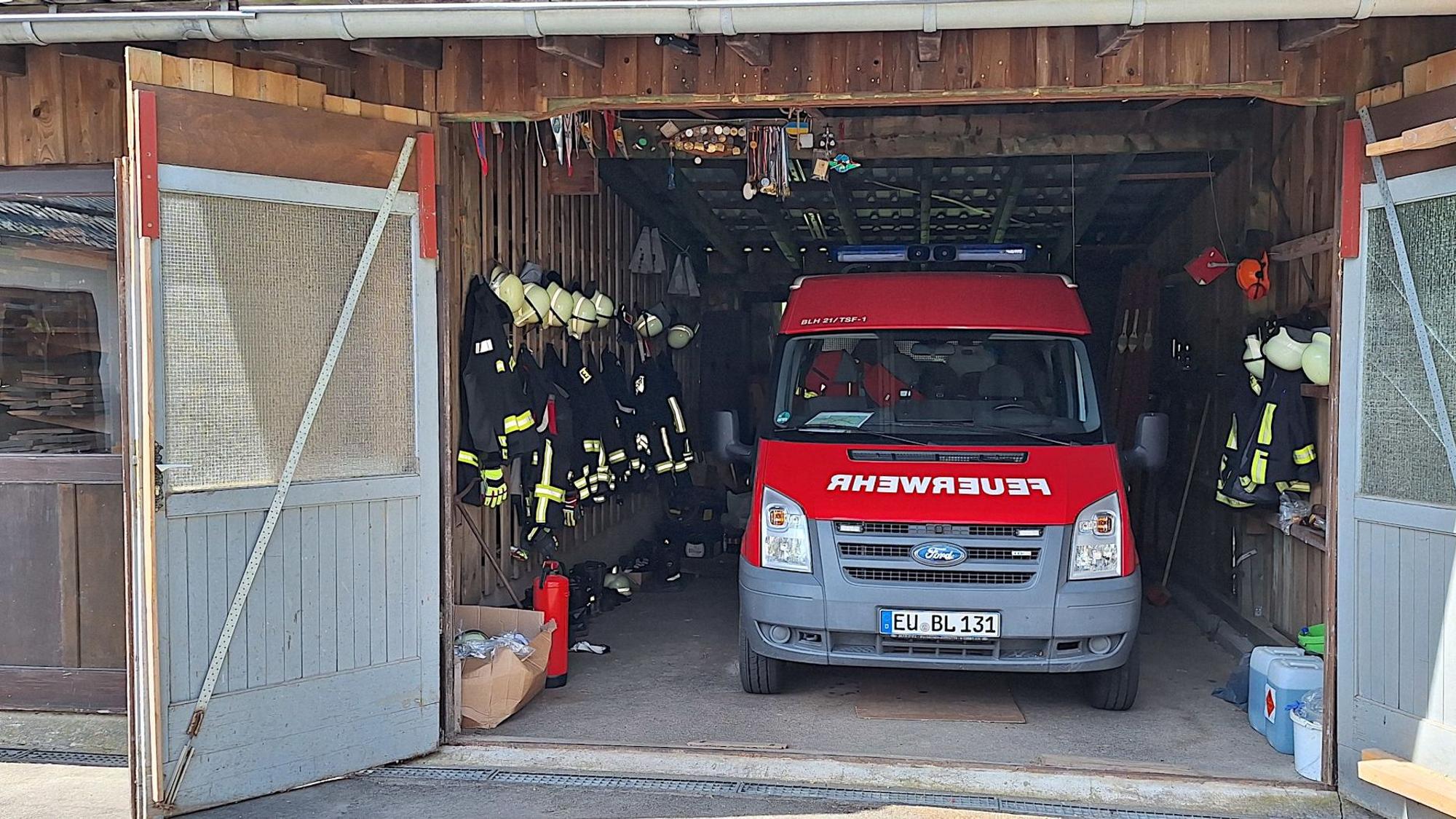 Ein rotes Feuerwehrauto steht in einm Holzschuppen. Links und rechts daneben hängt Feuerwehrkleidung.