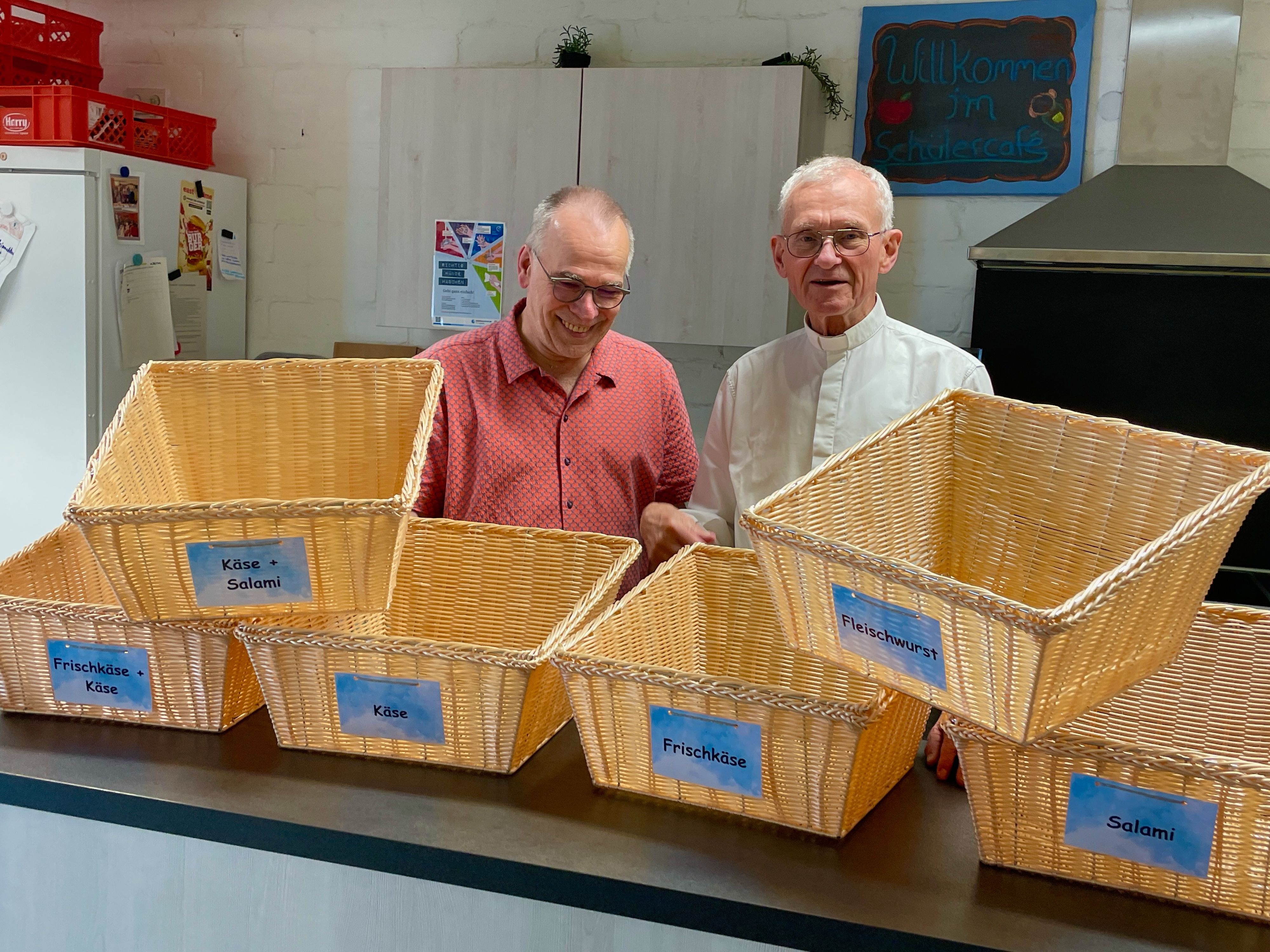 Zwei Männer stehen hinter einer Theke, auf der Brötchenkörbe mit verschiedenen Beschriftungen stehen.