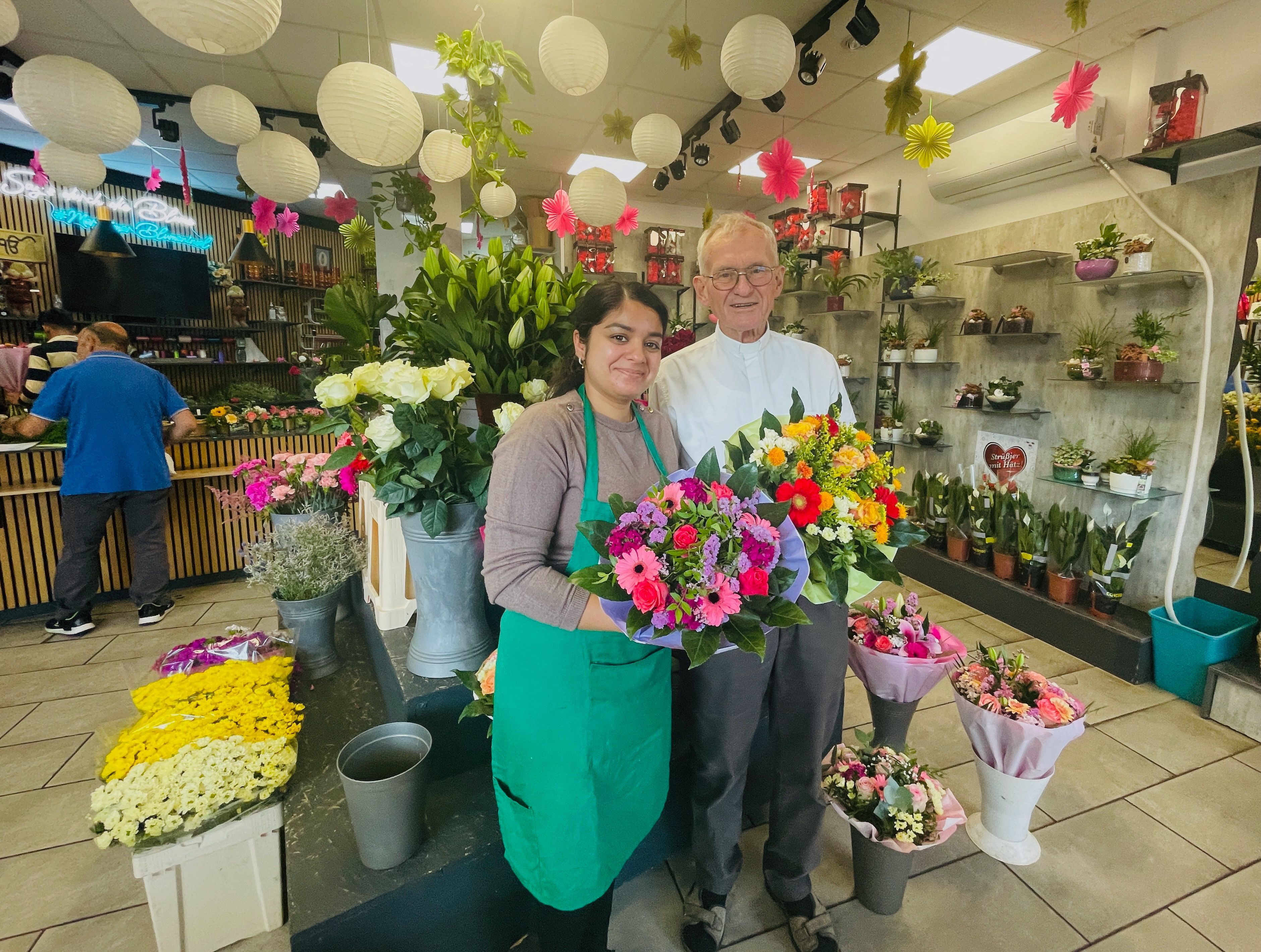 Eine junge Frau steht neben einem älteren Herrn in einem Blumenladen. Sie hält einen Strauß in der Hand.
