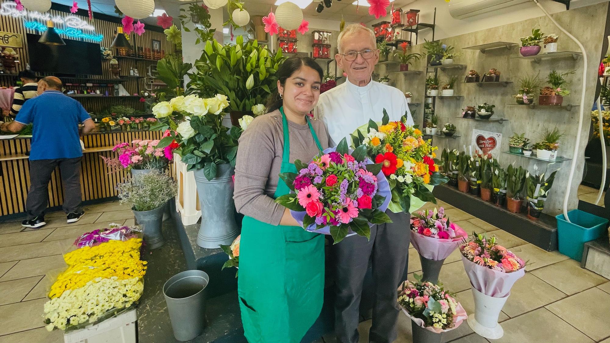 Eine junge Frau steht neben einem älteren Herrn in einem Blumenladen. Sie hält einen Strauß in der Hand.