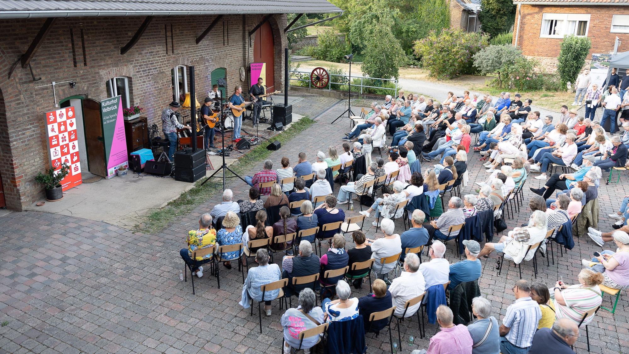 Die Musiker stehen unter einem Dachüberstand. Das Publikum sitzt in bogenförmigen Stuhlreihen.