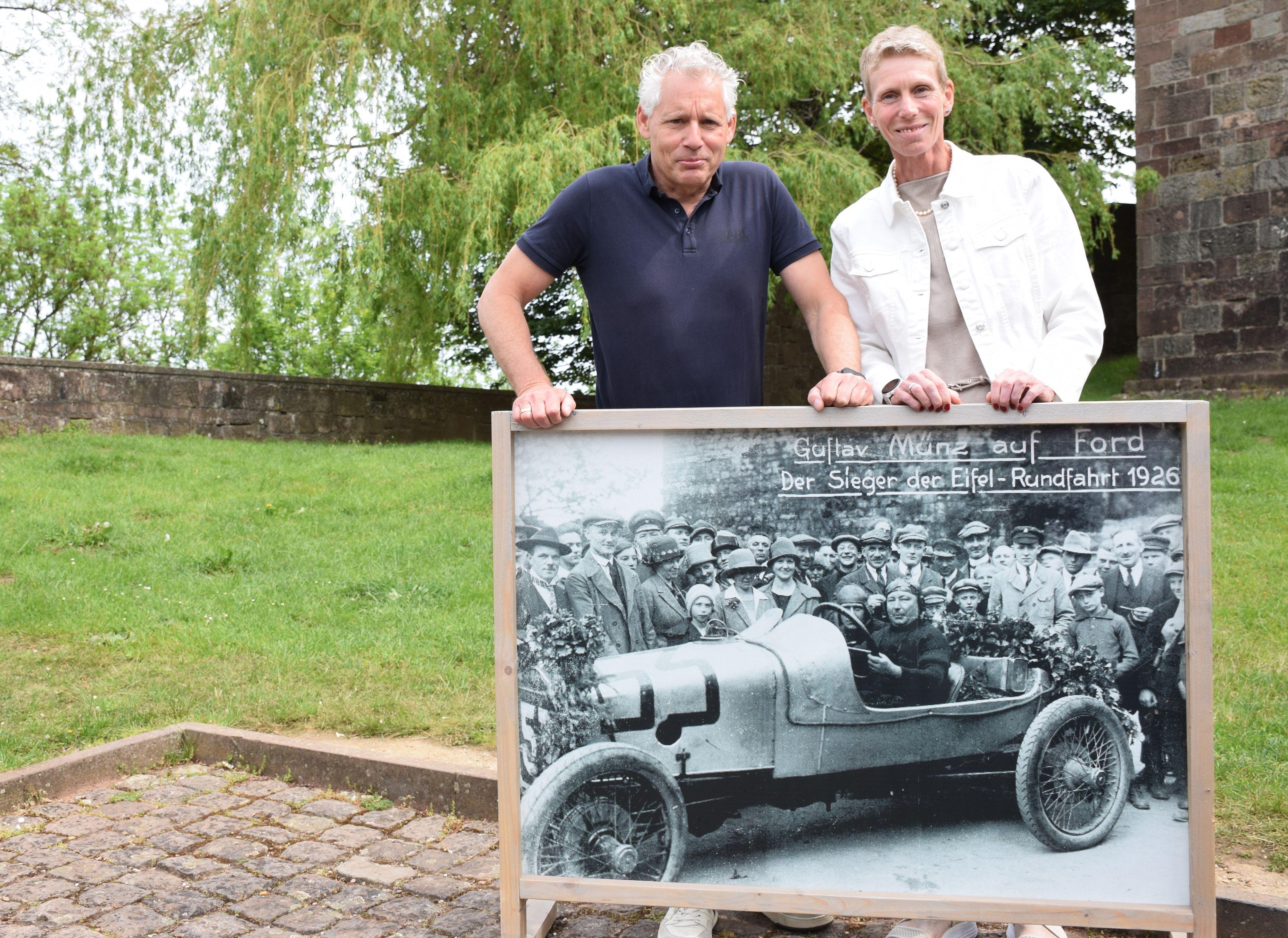 Georg Münz und Jette Bork-Wagenblast halten ein großformatiges Foto, das Gustav Münz als Sieger der Eifel-Rundfahrt 1926 in seinem Ford zeigt.
