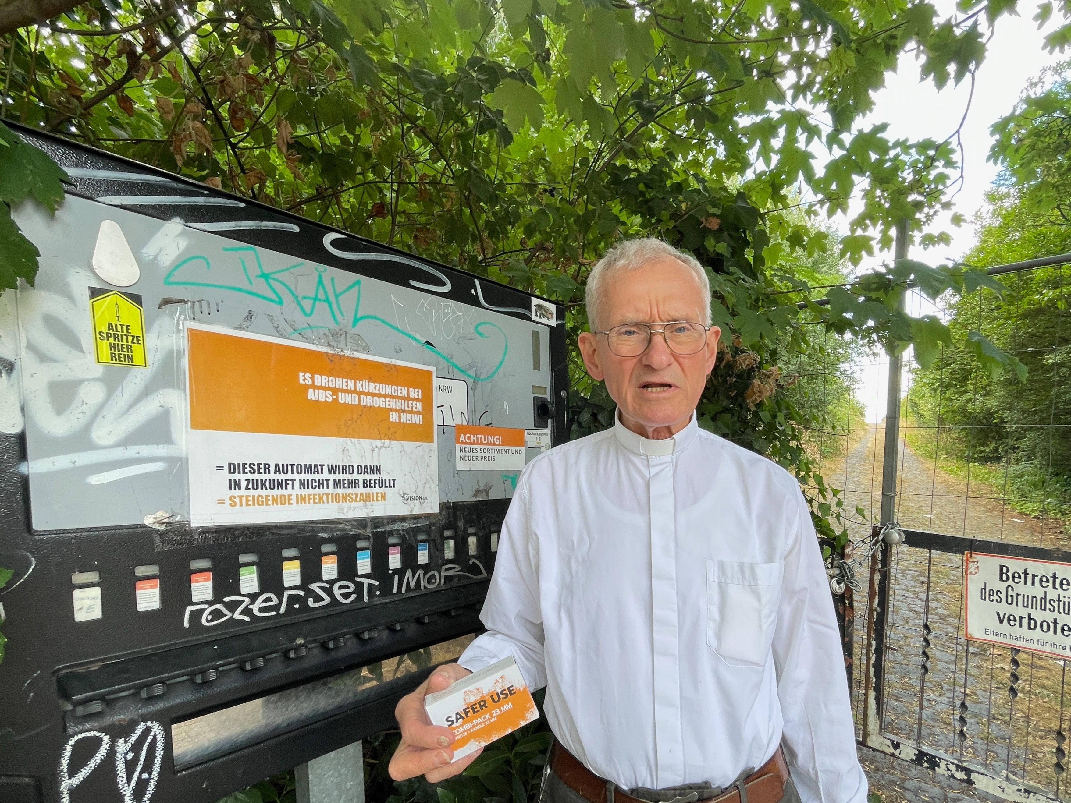 Ein Mann im weißen Hemd steht vor einem Spritzenautomat. Er hat ein Spritzen-Einweg-Set in der Hand.