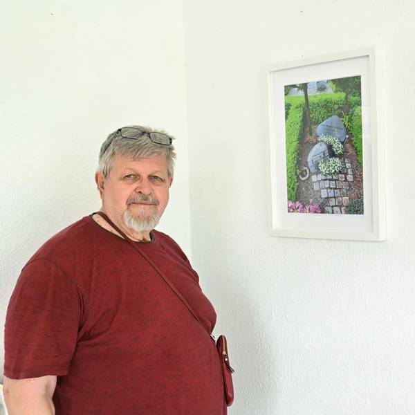 Ein bärtiger Mann in rotem T-Shirt steht neben zwei Fotografien, die gerahmt an der Wand hängen.
