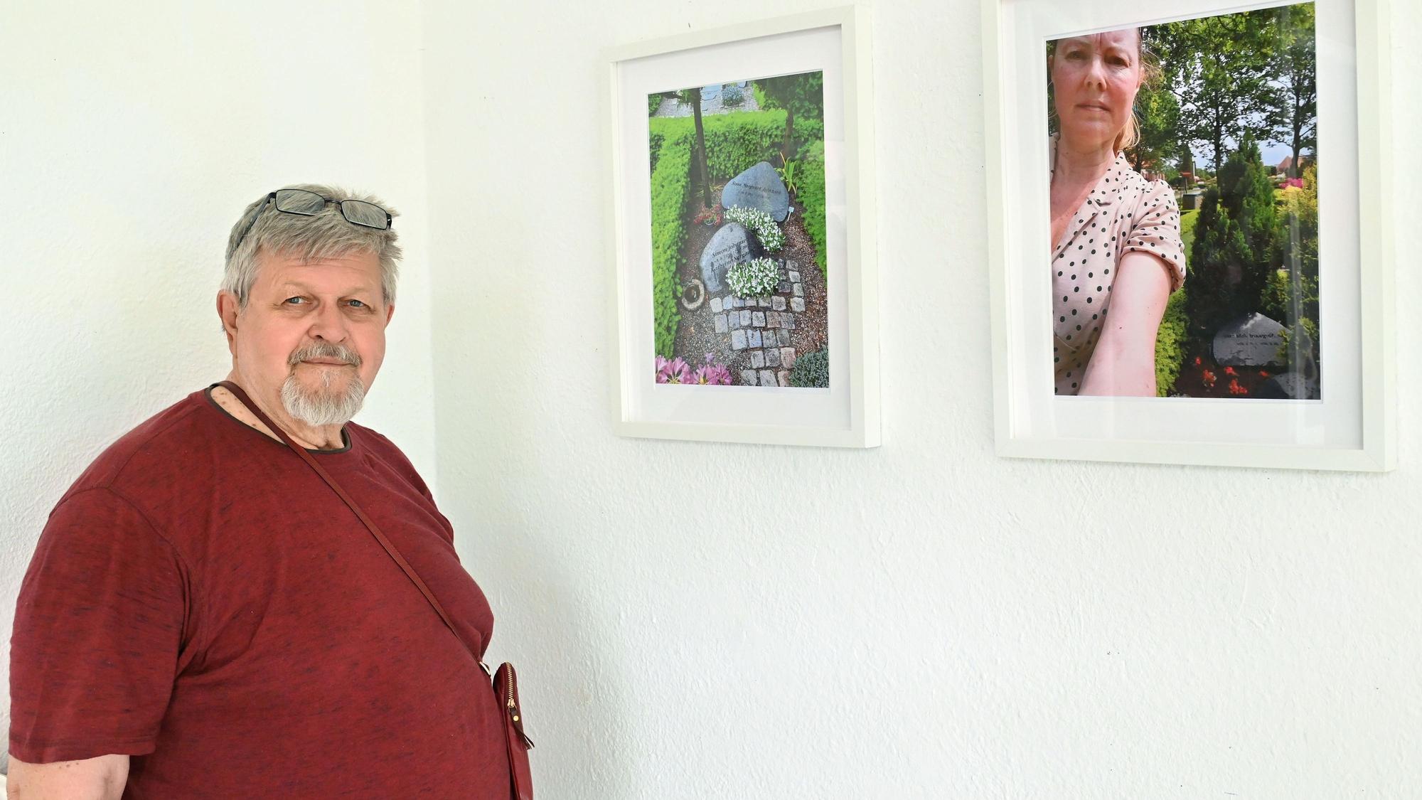 Ein bärtiger Mann in rotem T-Shirt steht neben zwei Fotografien, die gerahmt an der Wand hängen.