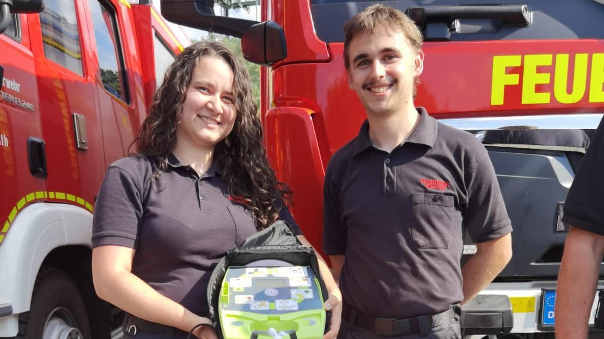 Zu sehen sind eine Feuerwehrfrau und ein Feuerwehrmann nebeneinander und sie lächeln. Sie hält einen Defibrillator in der Hand.