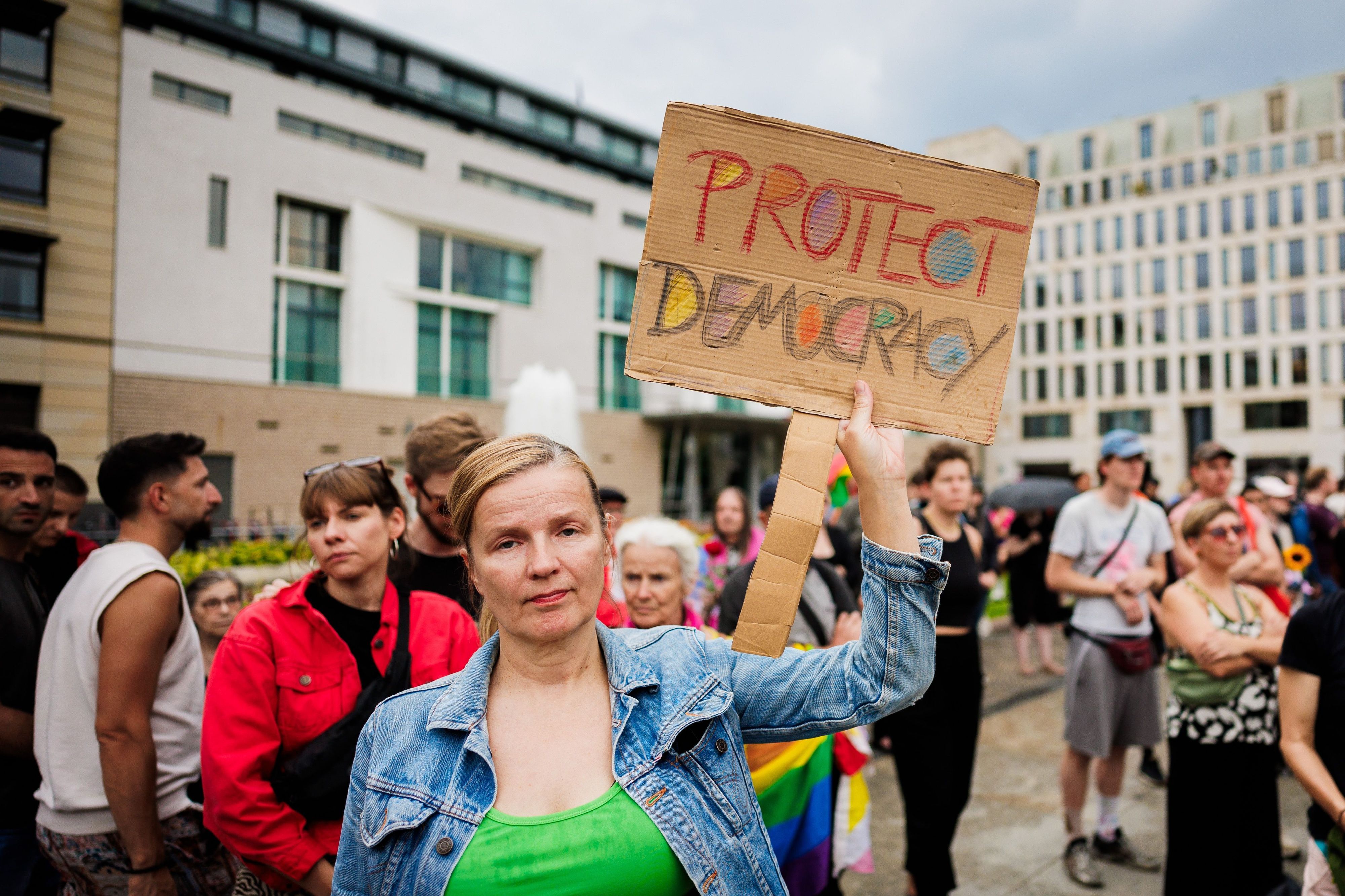 Menschen trauern und demonstrieren vor dem Brandenburger Tor. Eine Person hält dabei ein Schild mit der Aufschrift "Protect Democracy" (Schützt die Demokratie).