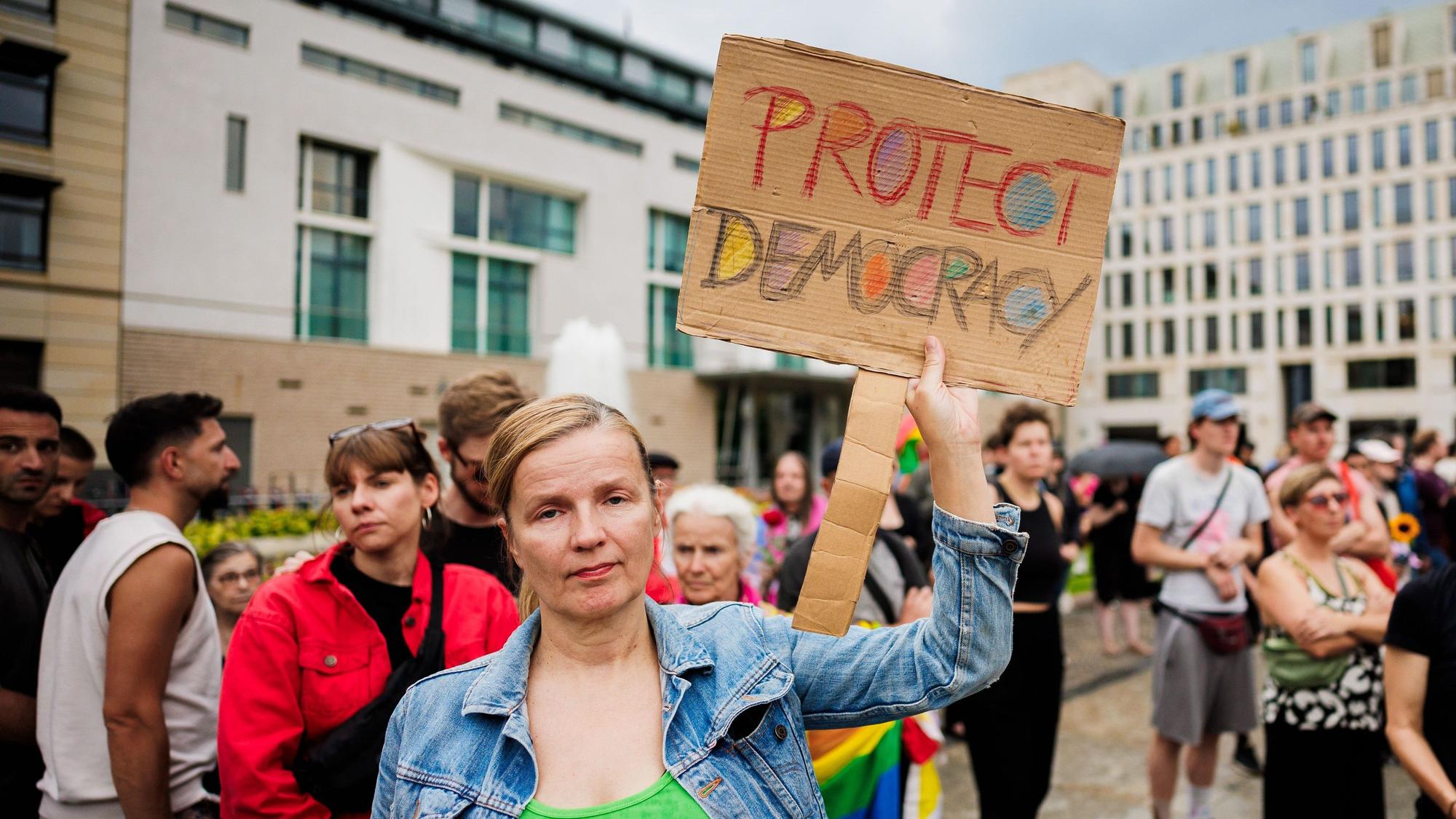 Menschen trauern und demonstrieren vor dem Brandenburger Tor. Eine Person hält dabei ein Schild mit der Aufschrift "Protect Democracy" (Schützt die Demokratie).