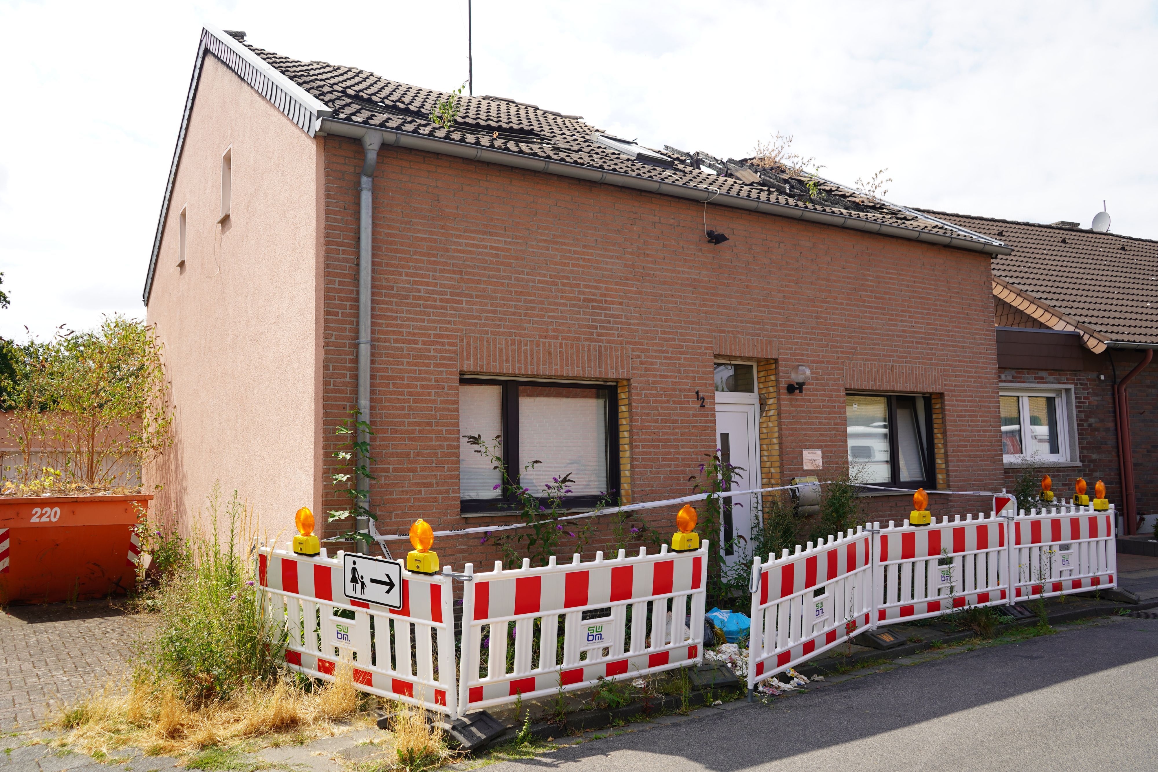 Das Bild zeigt ein Haus mit abgebranntem Dachstuhl. Davor ist der Gehweg abgesperrt.