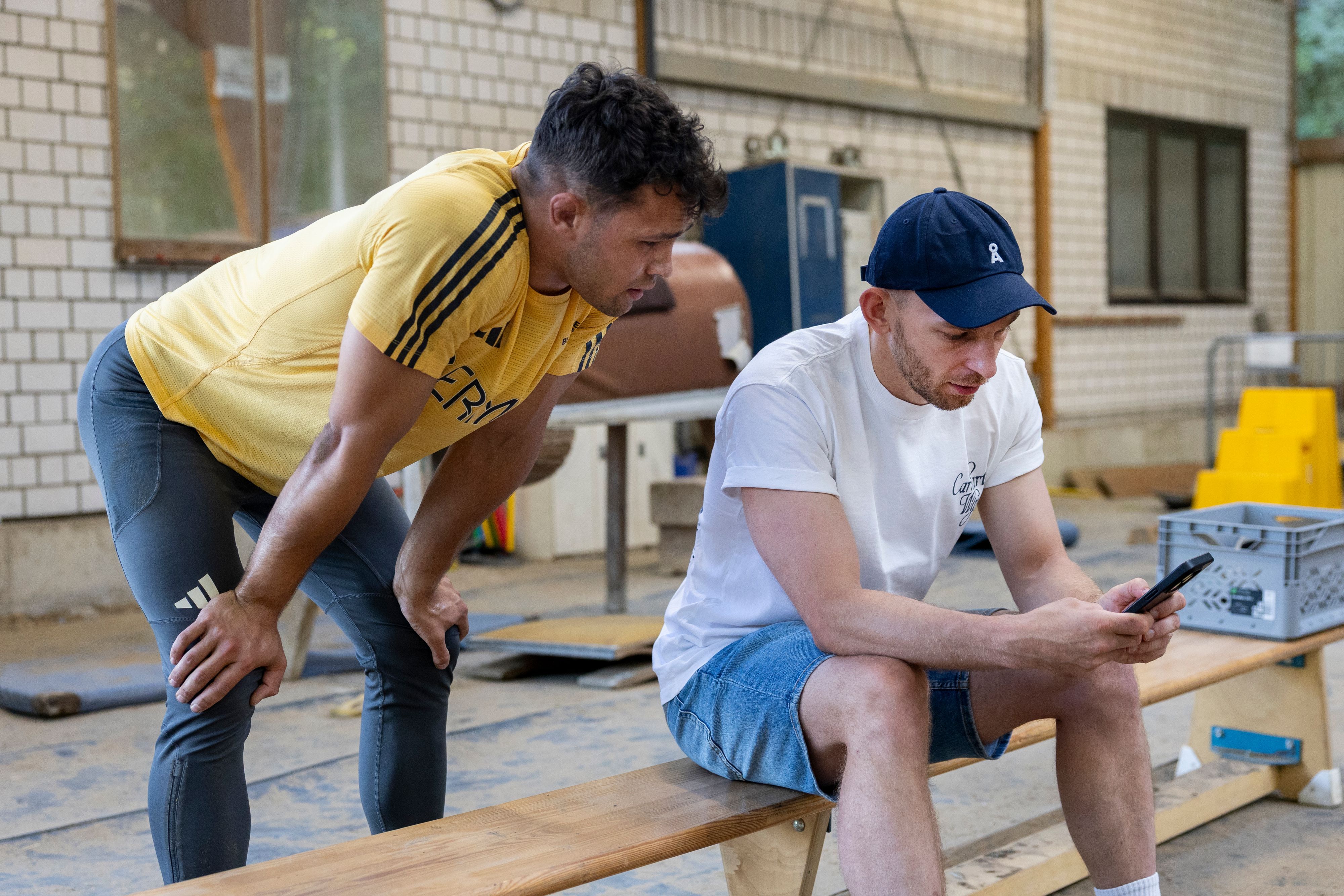 Ein junger Mann in Sportsachen steht hinter einem anderen Mann, der ihm auf einer Turnbank sitzend auf dem Handy Szenen seiner Kür-Übung zeigt.