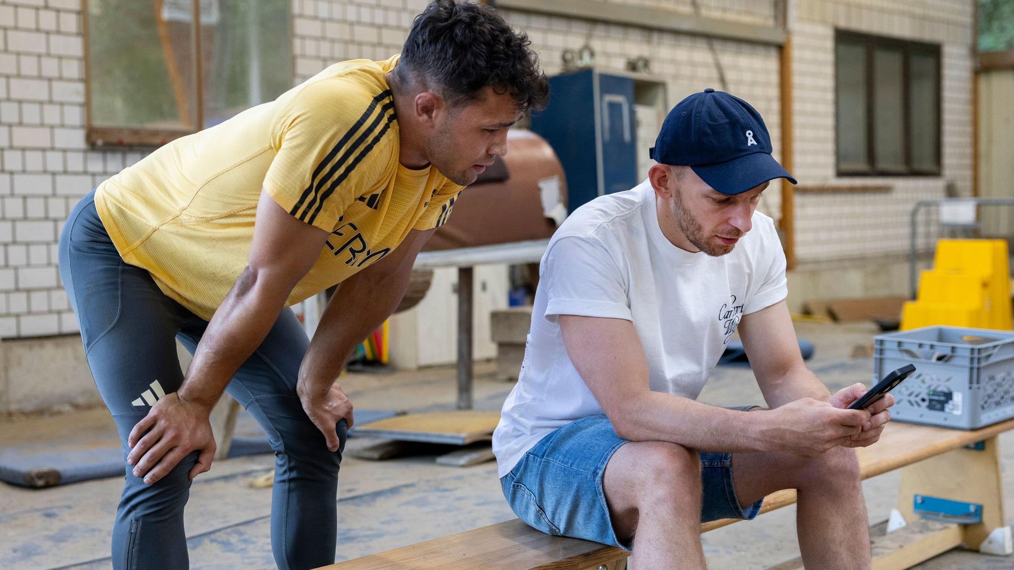 Ein junger Mann in Sportsachen steht hinter einem anderen Mann, der ihm auf einer Turnbank sitzend auf dem Handy Szenen seiner Kür-Übung zeigt.