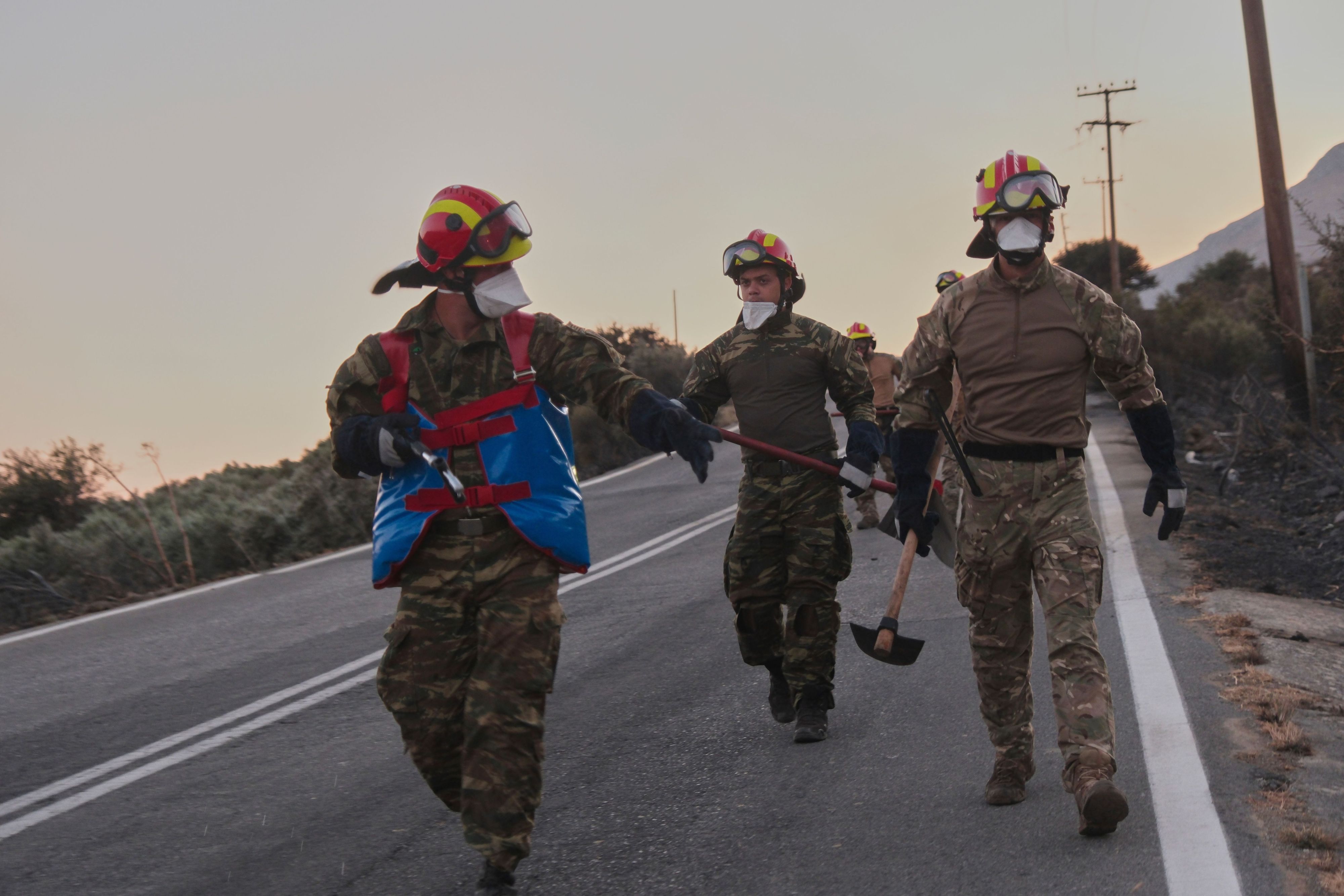 Feuerwehrleute gehen während eines Waldbrands in der Region Rethymno auf der südgriechischen Insel Kreta mit Werkzeugen eine Straße entlang.