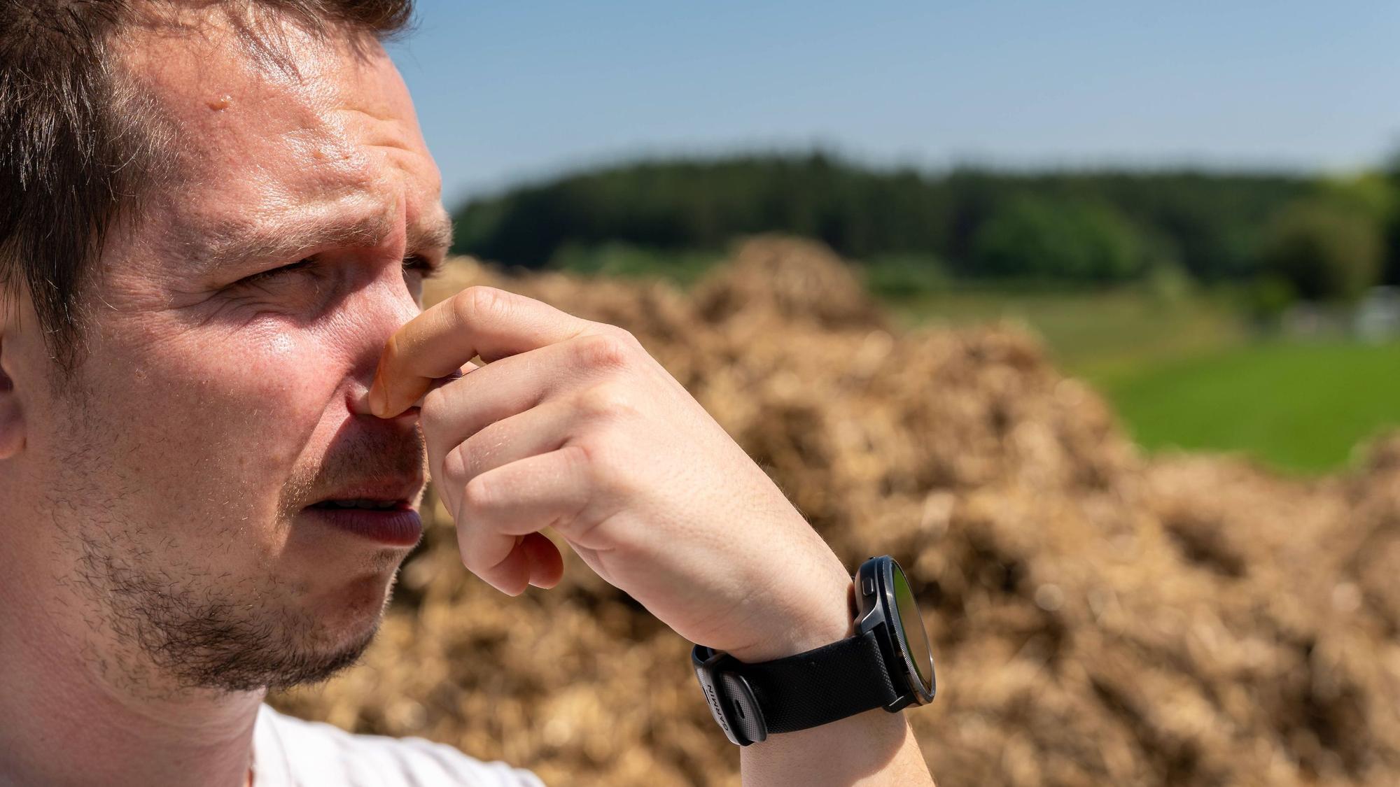 Ein junger Mann hält sich vor einem Misthaufen auf dem Bauernhof die Nase zu bei heißen Temperaturen.