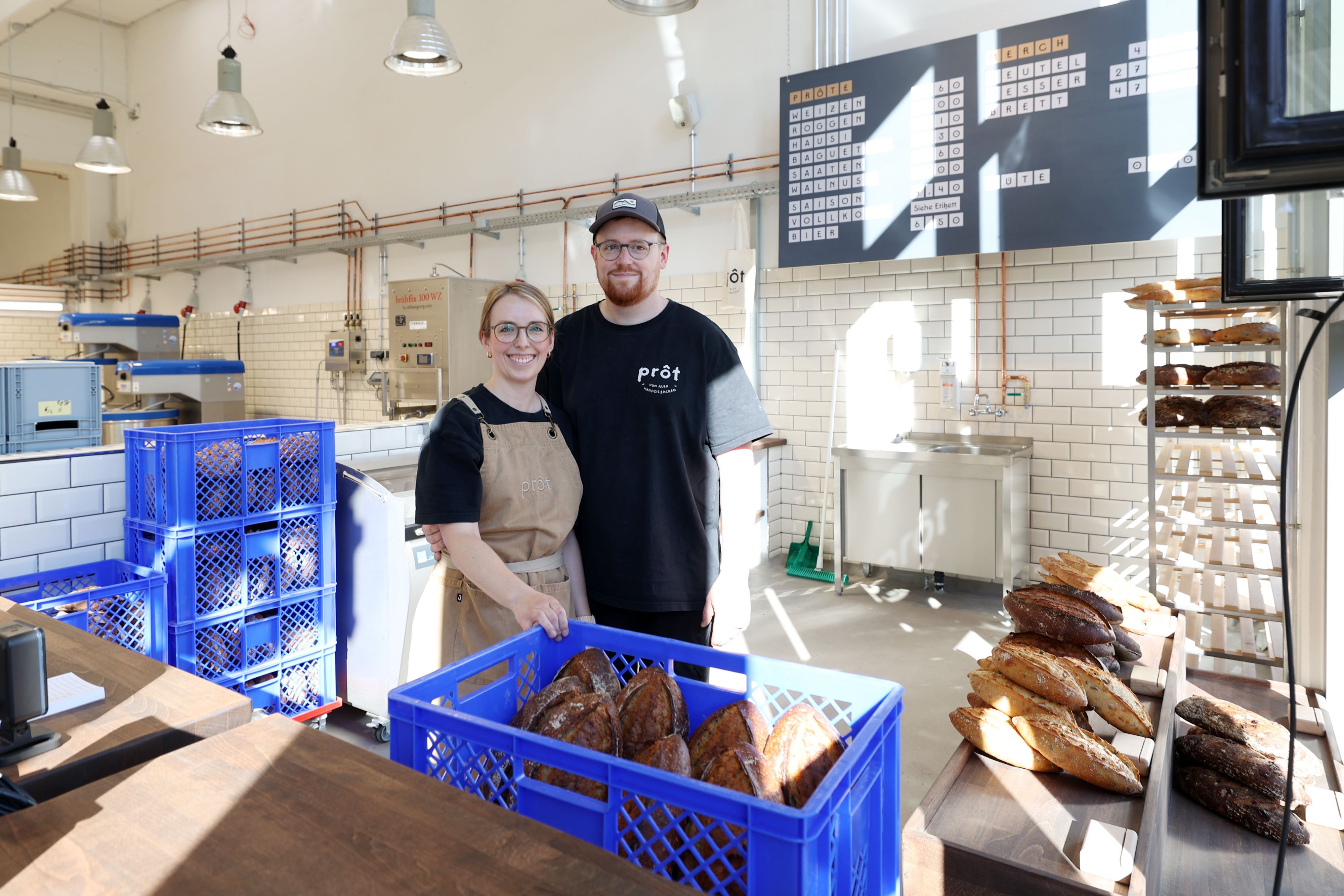 Eine Frau mit Zopf, Brille und Schürze steht neben einem Mann mit Cap und Bart. Sie stehen in einer großen Halle hinter einer Verkaufstheke, vor und neben sich sind Kisten mit Broten,
