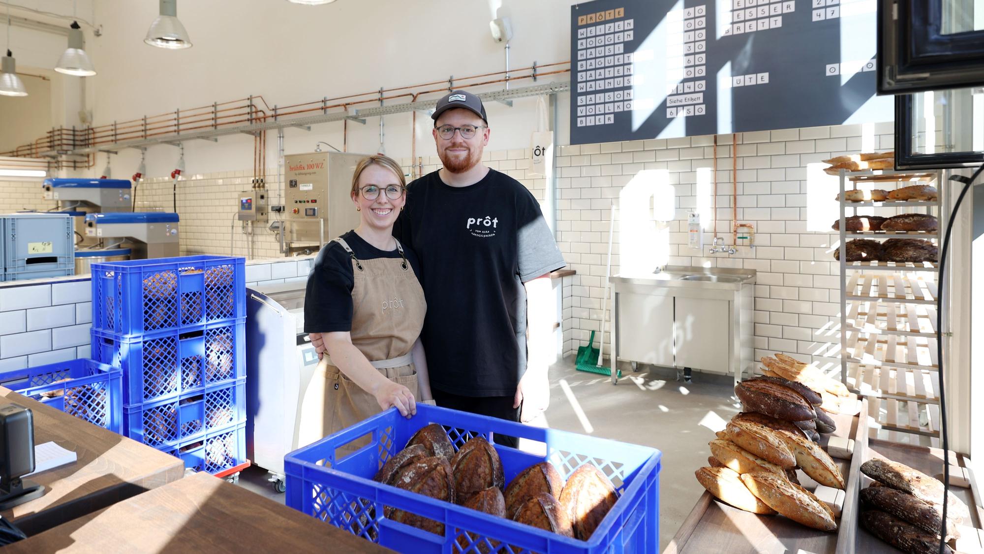Eine Frau mit Zopf, Brille und Schürze steht neben einem Mann mit Cap und Bart. Sie stehen in einer großen Halle hinter einer Verkaufstheke, vor und neben sich sind Kisten mit Broten,