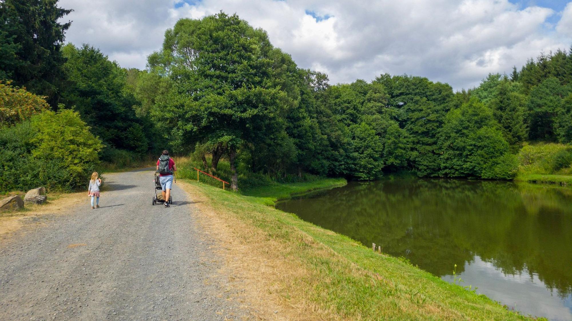 Ein Mann schiebt einen Kinderwagen über einen Weg neben einem See, links von ihm läuft ein Mädchen.
