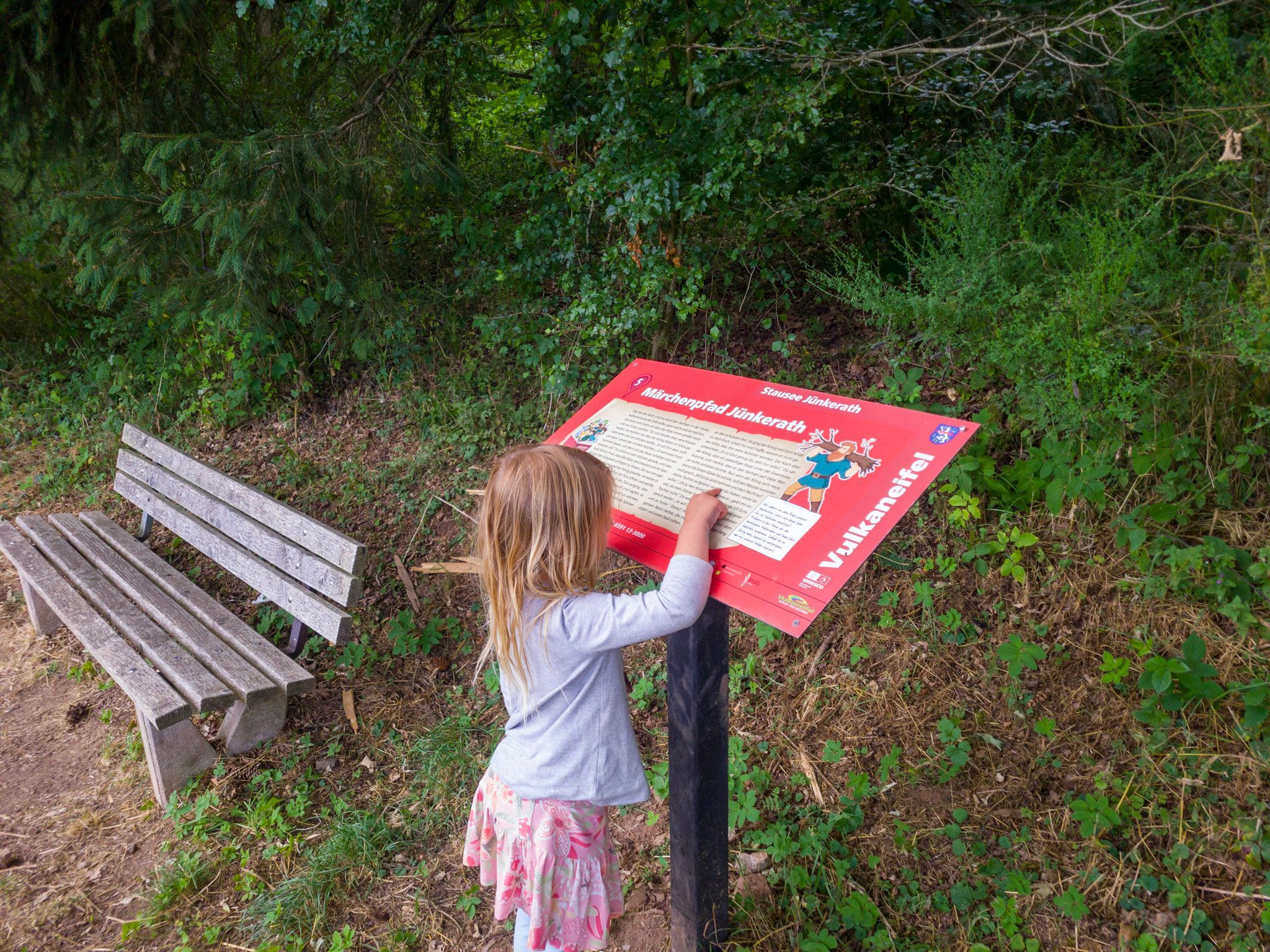 Ein Mädchen zeigt auf ein rotes Schild, das neben einer Bank am Wegesrand steht.