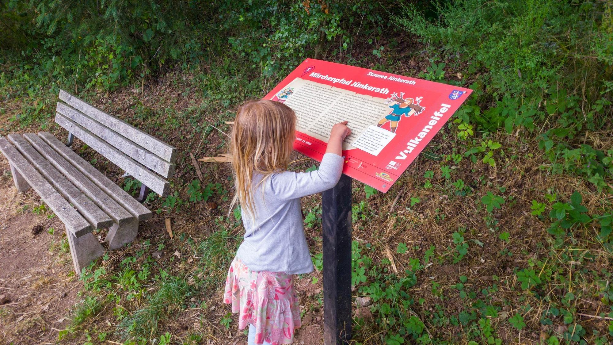 Ein Mädchen zeigt auf ein rotes Schild, das neben einer Bank am Wegesrand steht.