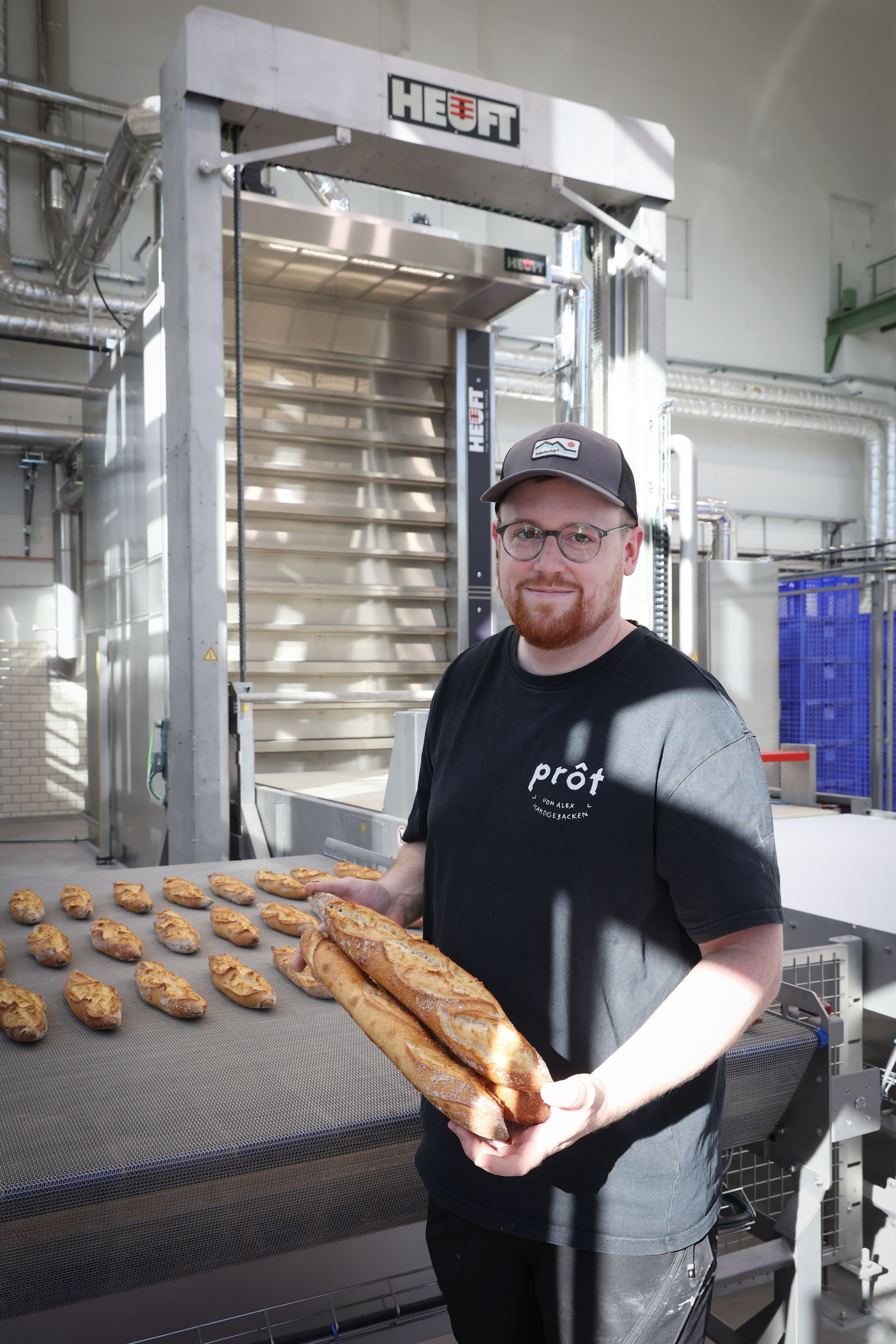 Ein Mann mit Cap, Brille und Bart steht vor der Ausgaberampe eines Ofens, auf der Dutzende Baguettes liegen. Zwei hält er in der Hand.