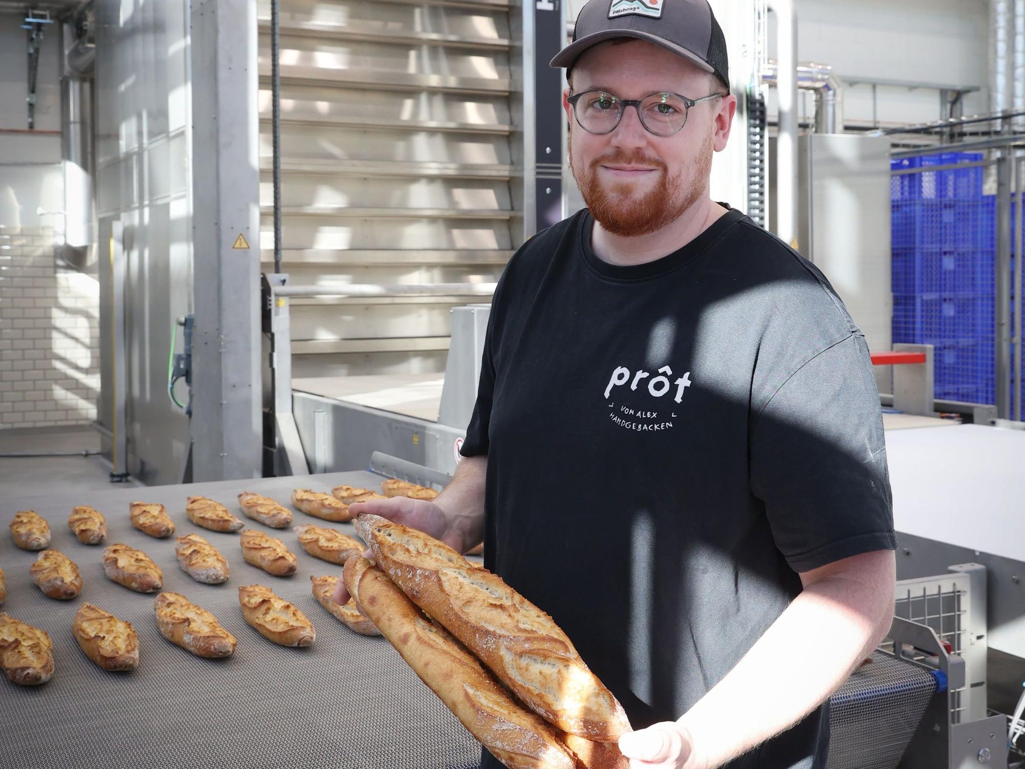 Ein Mann mit Cap, Brille und Bart steht vor der Ausgaberampe eines Ofens, auf der Dutzende Baguettes liegen. Zwei hält er in der Hand.