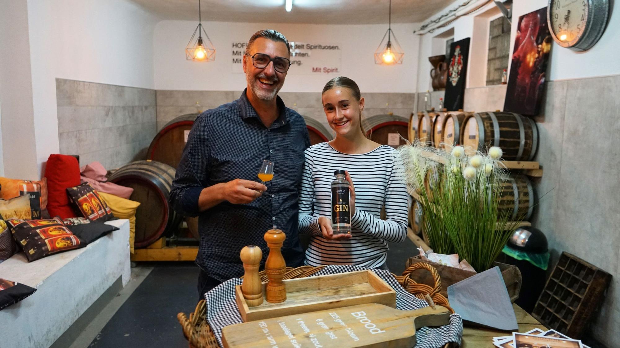 Ein Mann und eine junge Frau in einem Keller mit Weinfässern im Hintergrund, sie halten eine Ginflasche und ein Glas in den Händen