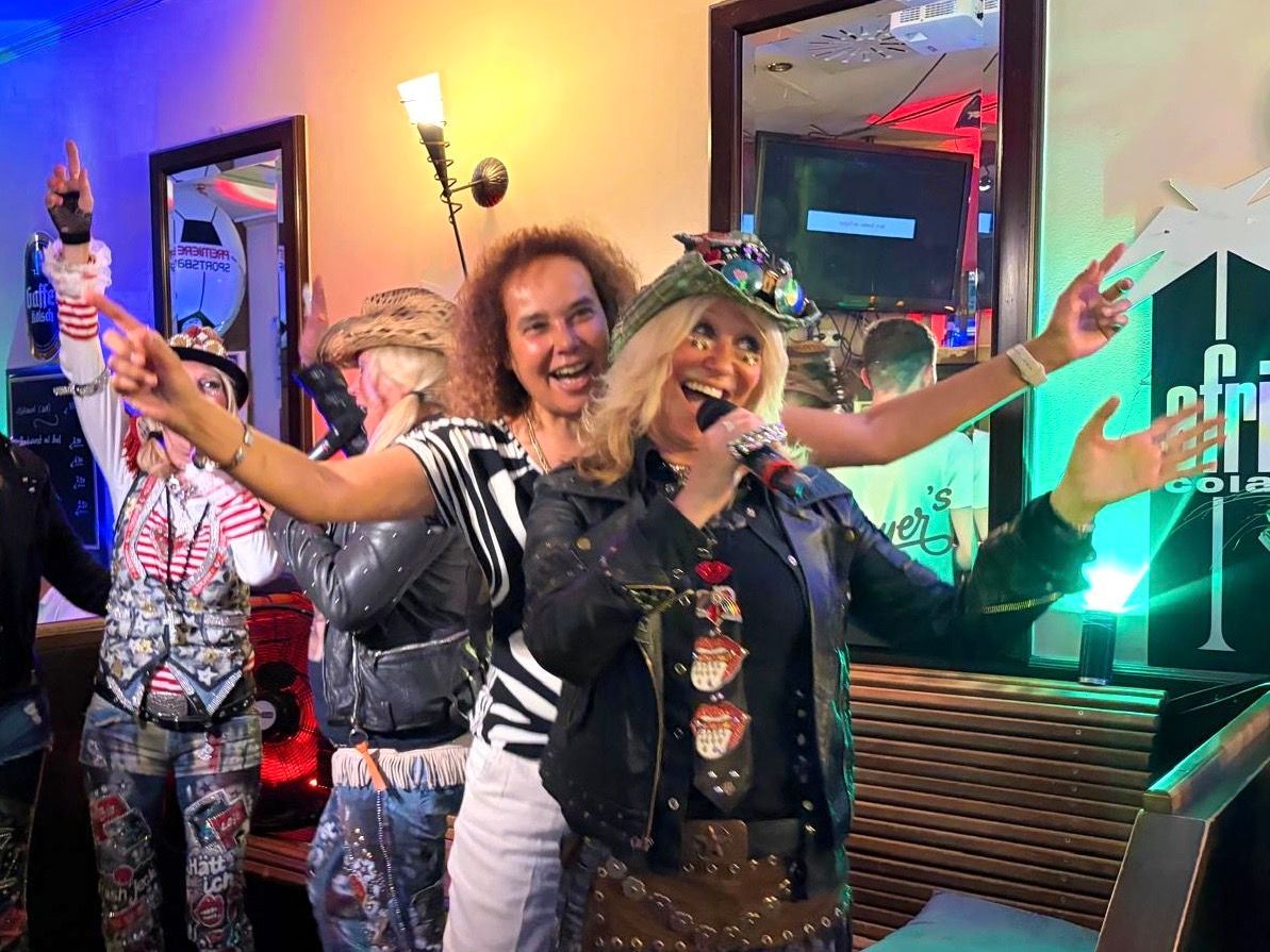 Frauen in Partykleidung singen gut gelaunt in ein Mikrofon, während sie sich in einer Gaststätte befinden.
