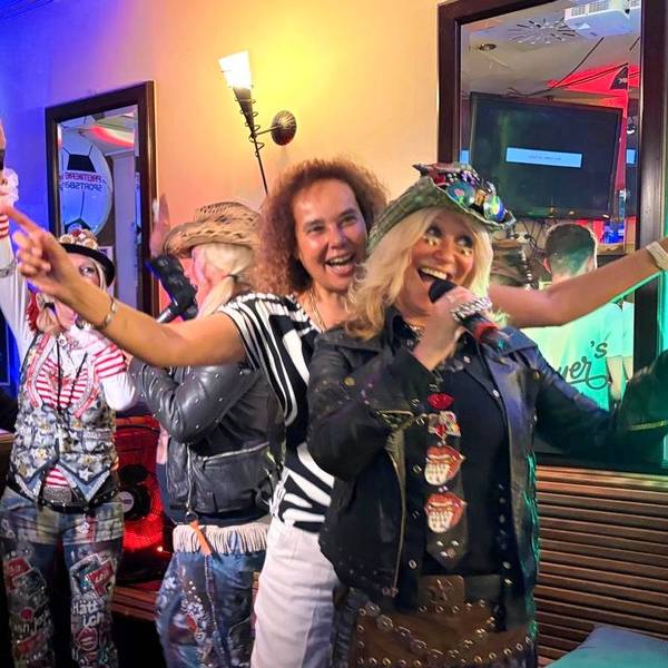 Frauen in Partykleidung singen gut gelaunt in ein Mikrofon, während sie sich in einer Gaststätte befinden.