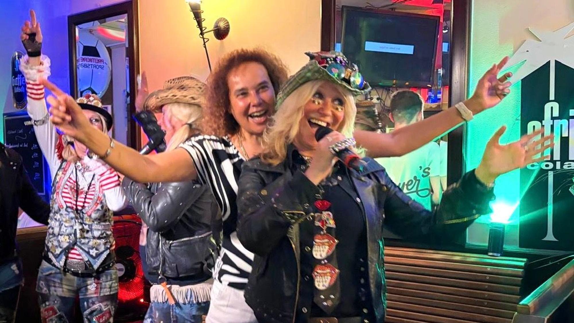 Frauen in Partykleidung singen gut gelaunt in ein Mikrofon, während sie sich in einer Gaststätte befinden.