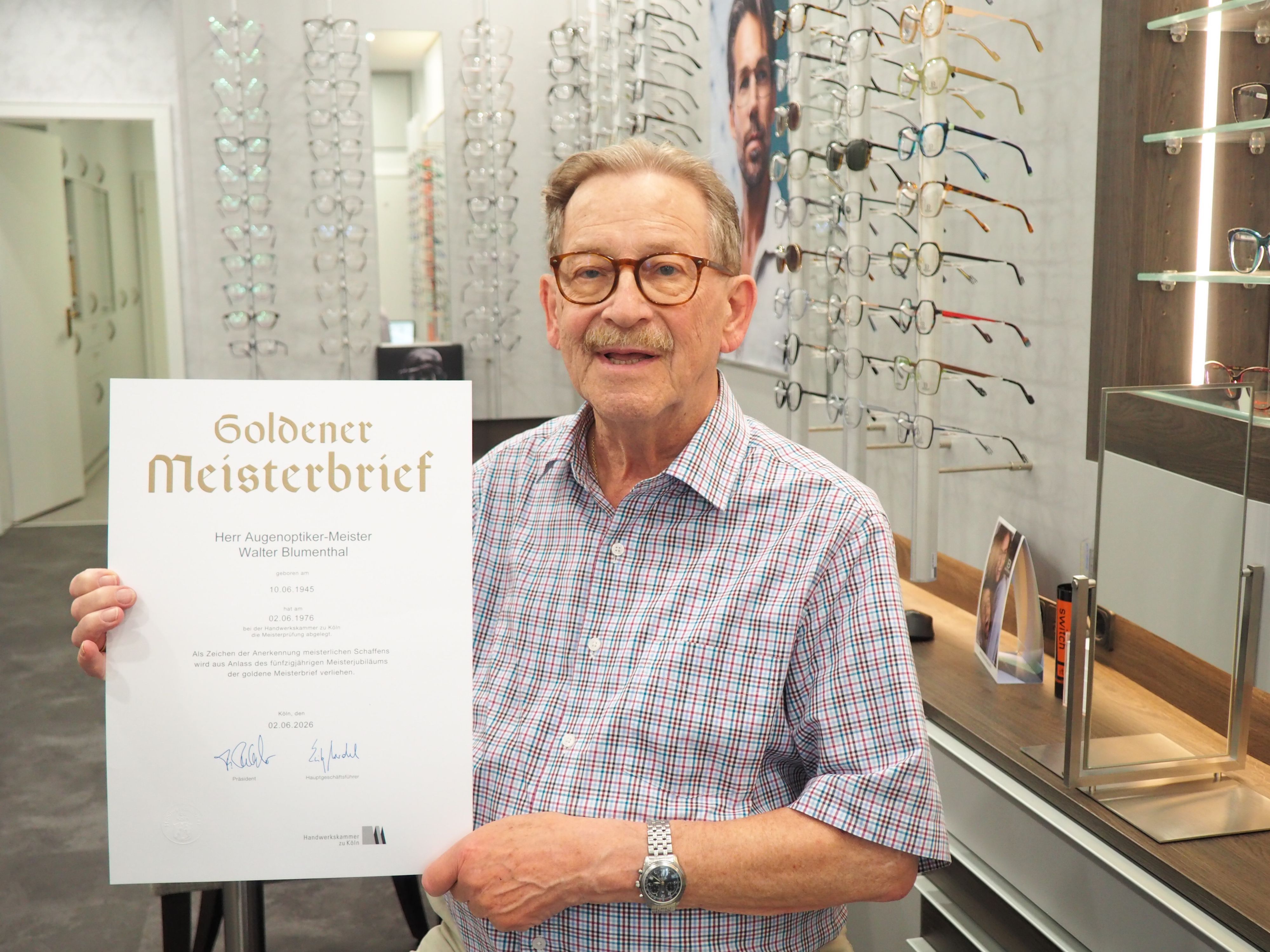 Walter Blumenthal zeigt seinen Goldenen  Meisterbrief, an den Wänden hängen Brillengestelle.