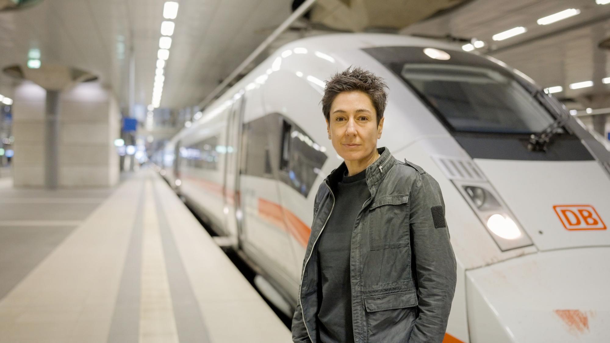 Dunja Hayali steht in einer Bahnhofshalle auf einem leeren Gleis vor einem ICE und schaut in die Kamera.