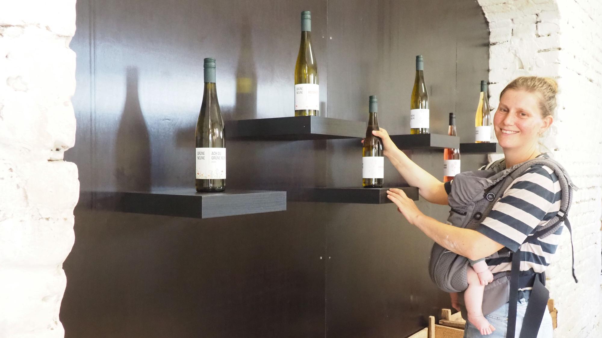 Eine Frau hält eine Flasche in einer Hand und steht vor einer Wand, an der einzelne Flaschen präsentiert werden.