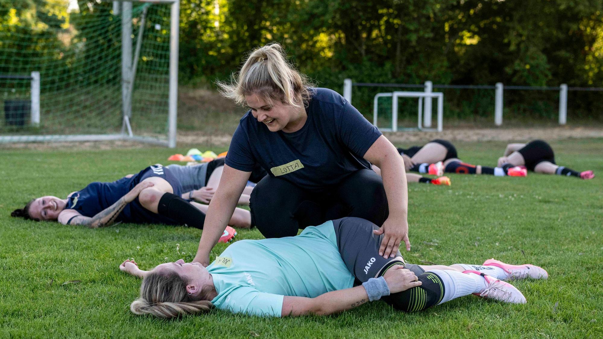Schäfer hilft einer Spielerin, die auf dem Rasen liegt, bei einer Dehnübung. Im Hintergrund führen weitere Sportlerinnen diese Übung aus.
